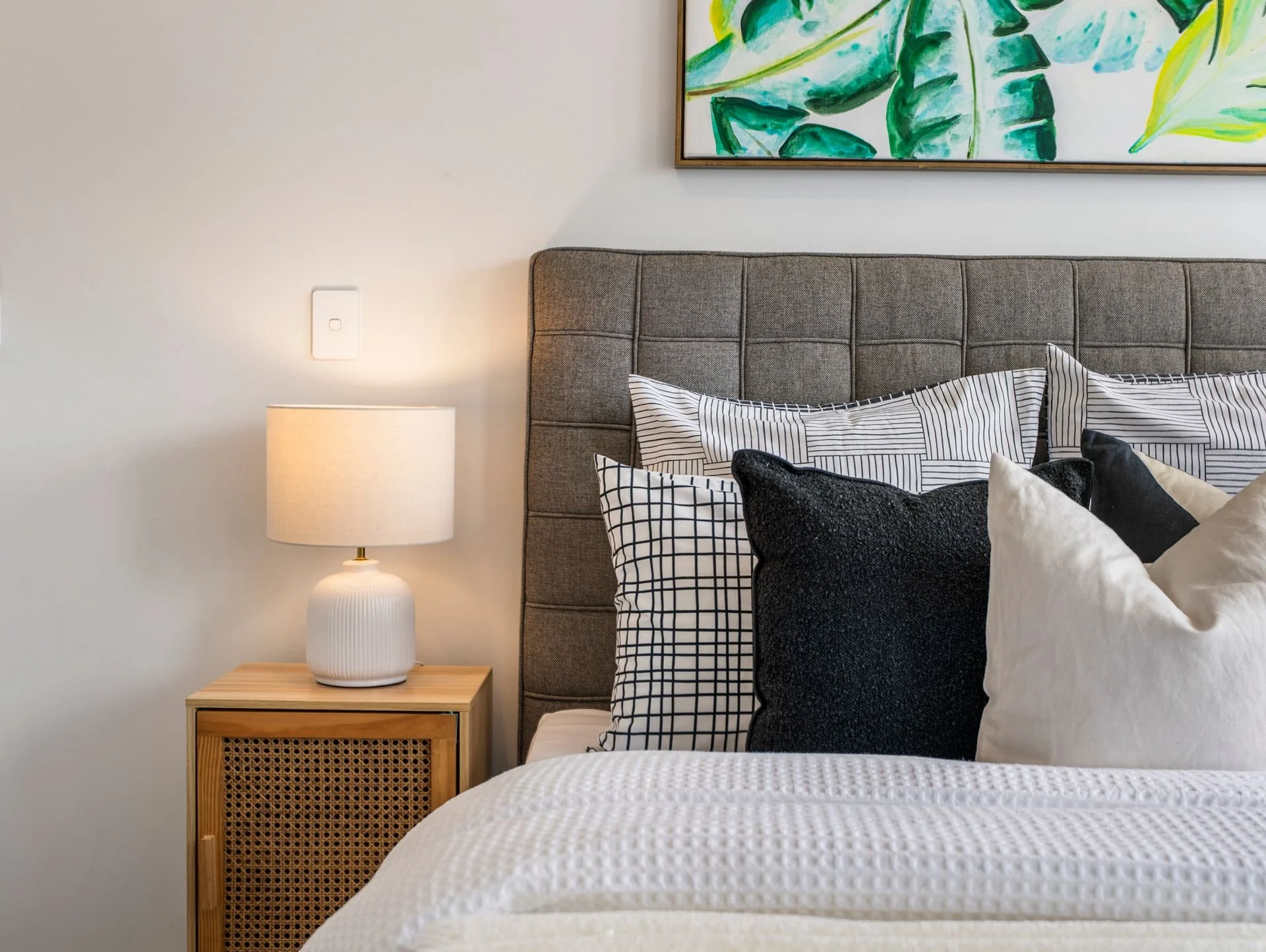 A bedroom with a gray upholstered headboard, featuring a variety of pillows in black, white, and striped patterns, next to a bedside table with a white textured lamp, a white wall with a switch, and a framed painting of green tropical leaves.