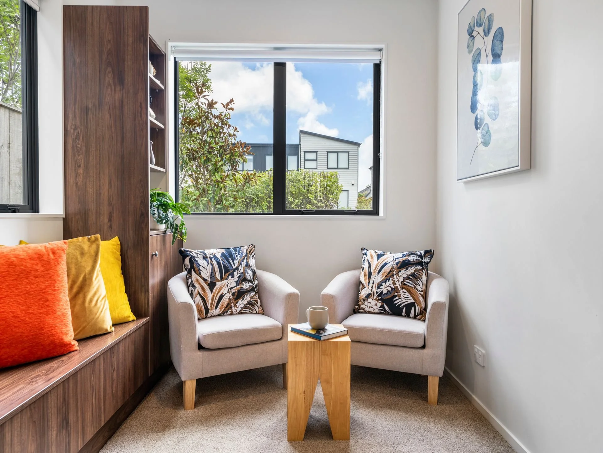 A small cozy room with two light-colored armchairs with patterned cushions, a small wooden side table with a mug and a book, a large window with a view of neighboring buildings and greenery, colorful cushions on a built-in wooden bench, a framed bota