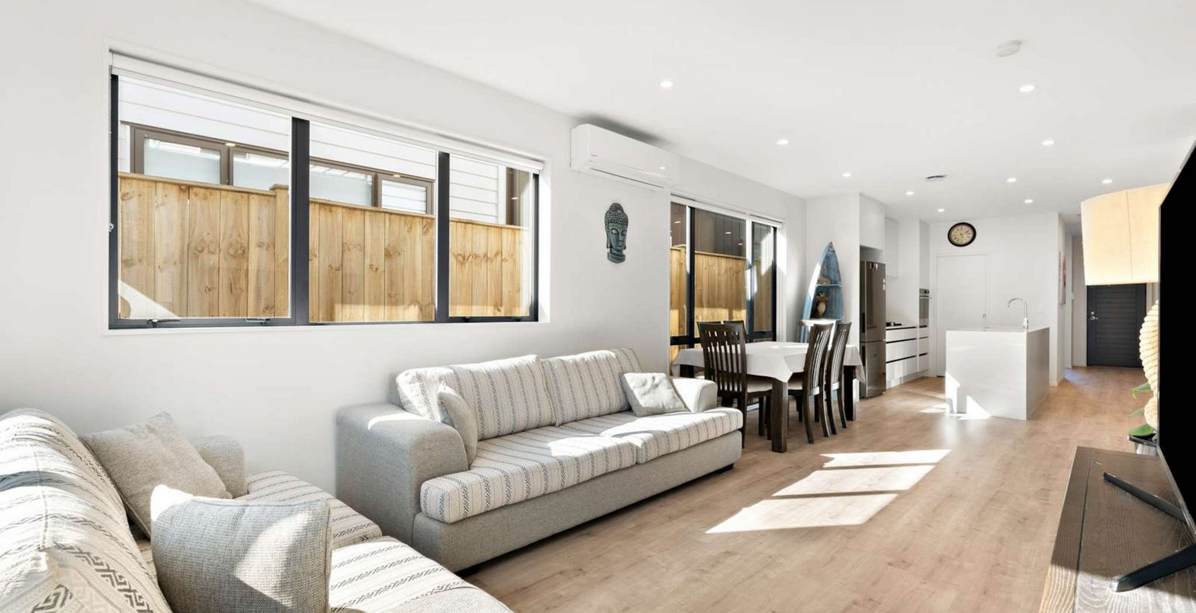 Bright living room and dining area with large windows, white walls, wooden flooring, a sofa, a dining table with chairs, and kitchen appliances in the background.