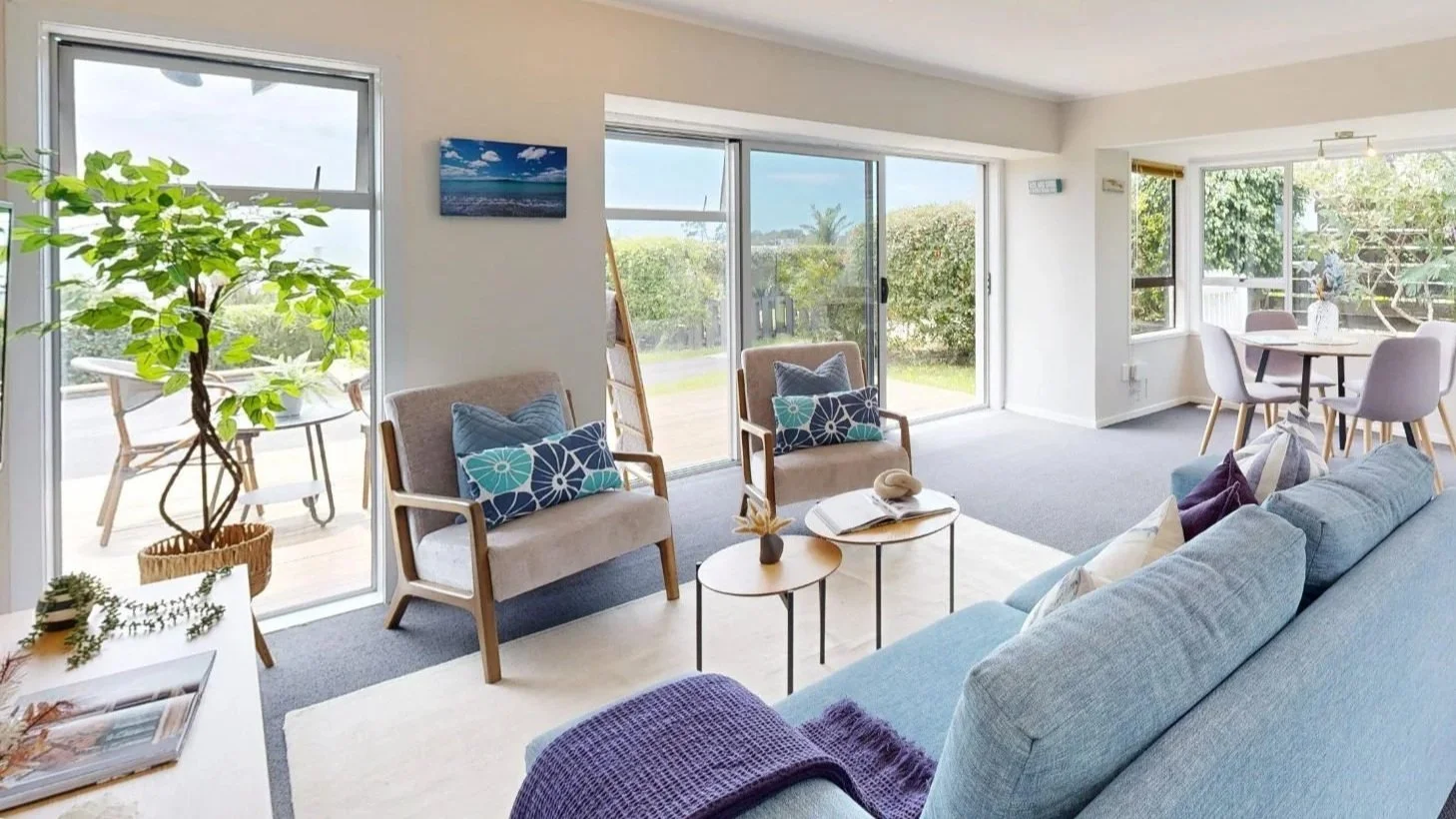 Living room with large windows, sliding glass doors, and patio furniture outside. Features a blue sofa with throw pillows, two beige armchairs with pillows, and a white coffee table with decorative items. There is a small table with plants and a large potted plant inside.