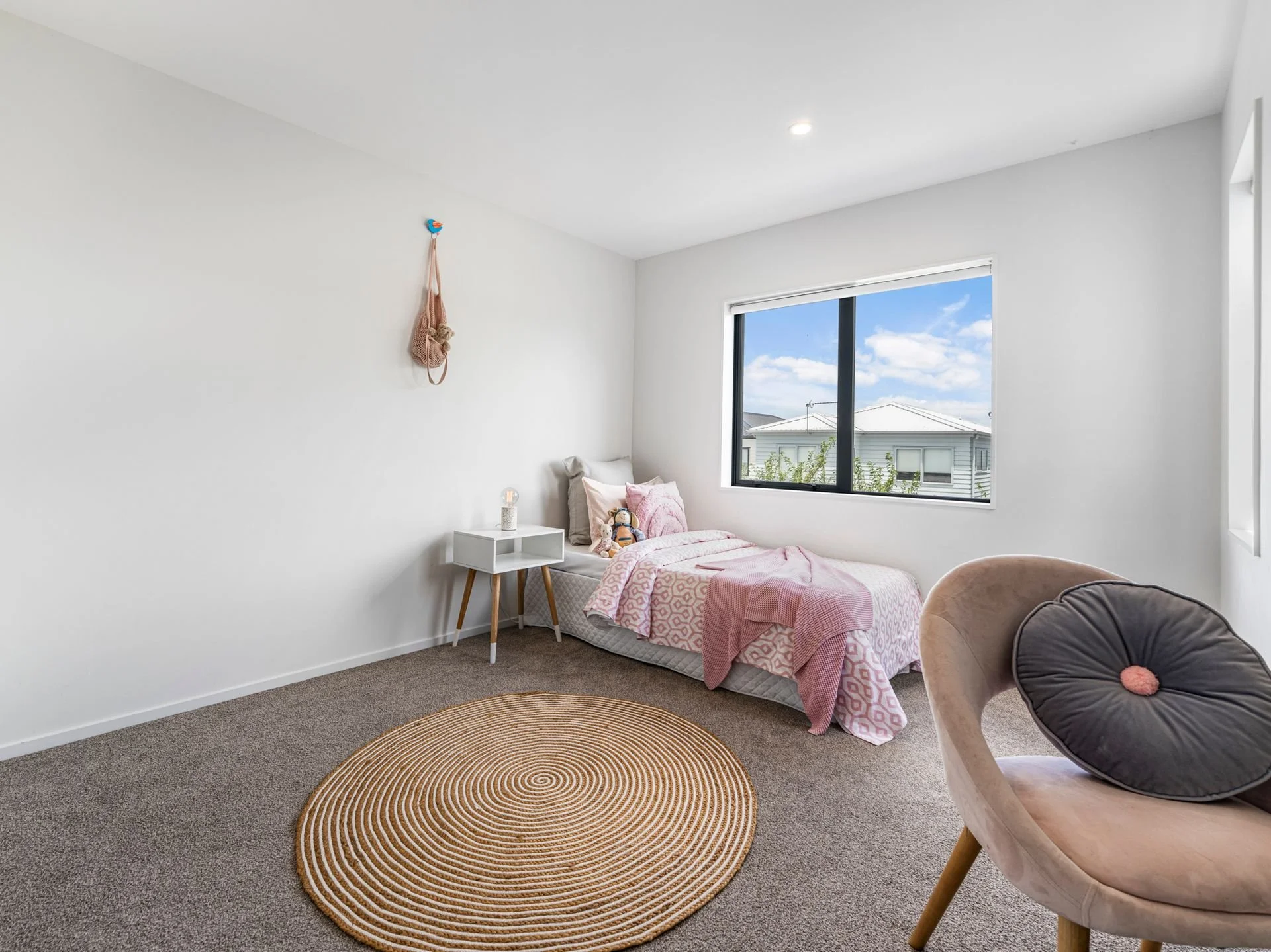 A minimalist child's bedroom with white walls, a single bed with pink bedding, a white side table, a tan circular rug, and a tan armchair with a dark gray pillow. There's a window showing a blue sky with clouds and neighboring houses.