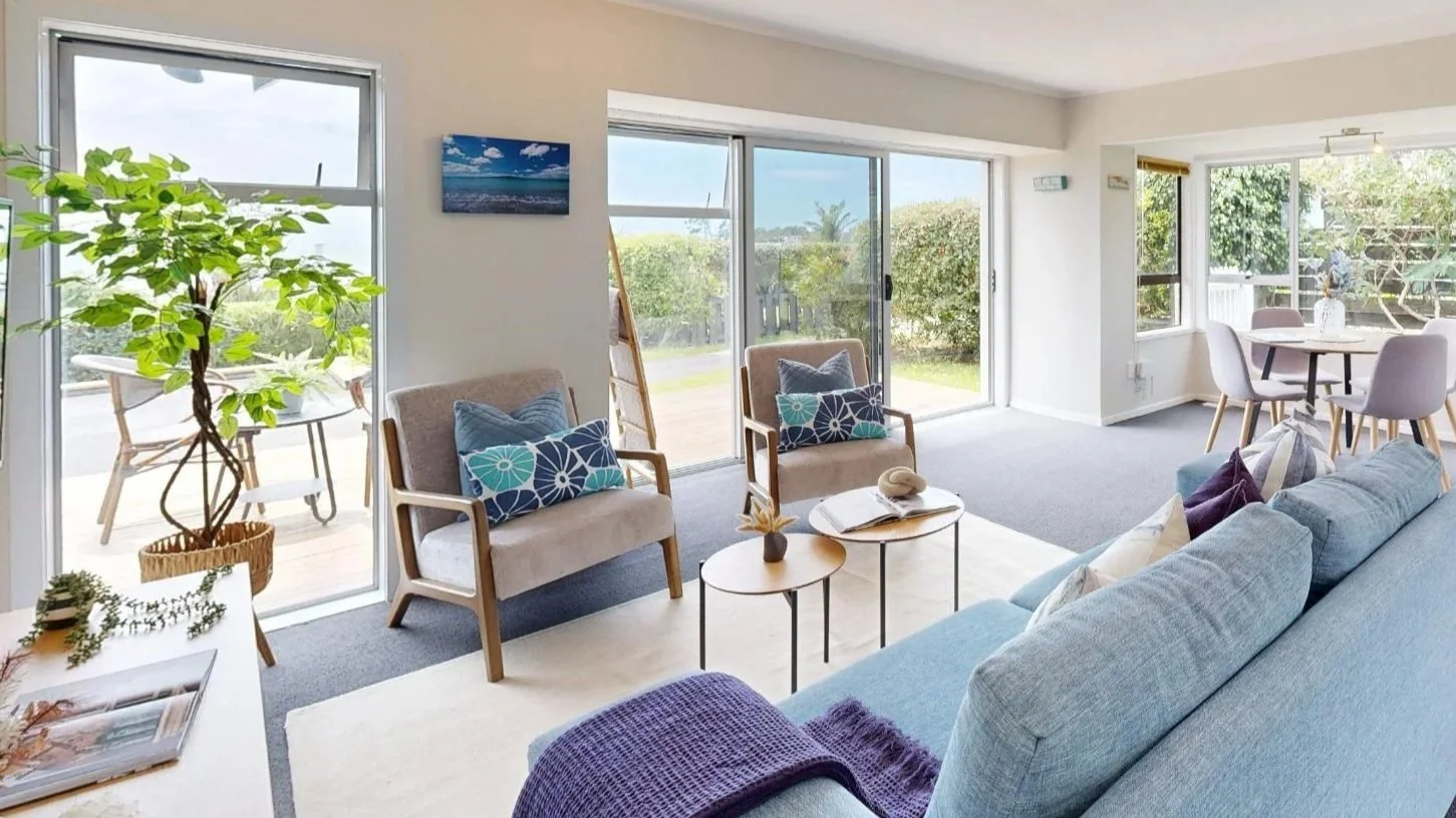 Bright living room with large windows, beige armchairs with blue and patterned cushions, a light blue sofa with pillows, a small round coffee table with a book and decorative items, a dining area with a round table and four chairs, outdoor patio with a table and chairs, greenery outside, and a potted plant inside.