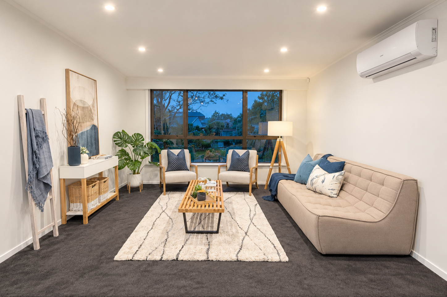 A cozy living room with a cream-colored sofa decorated with various pillows, a wooden side table with a small flower arrangement, a large floor lamp with a woven base, a window with gray curtains showing a garden outside, and a wall art piece depicting a beach scene with palm trees.