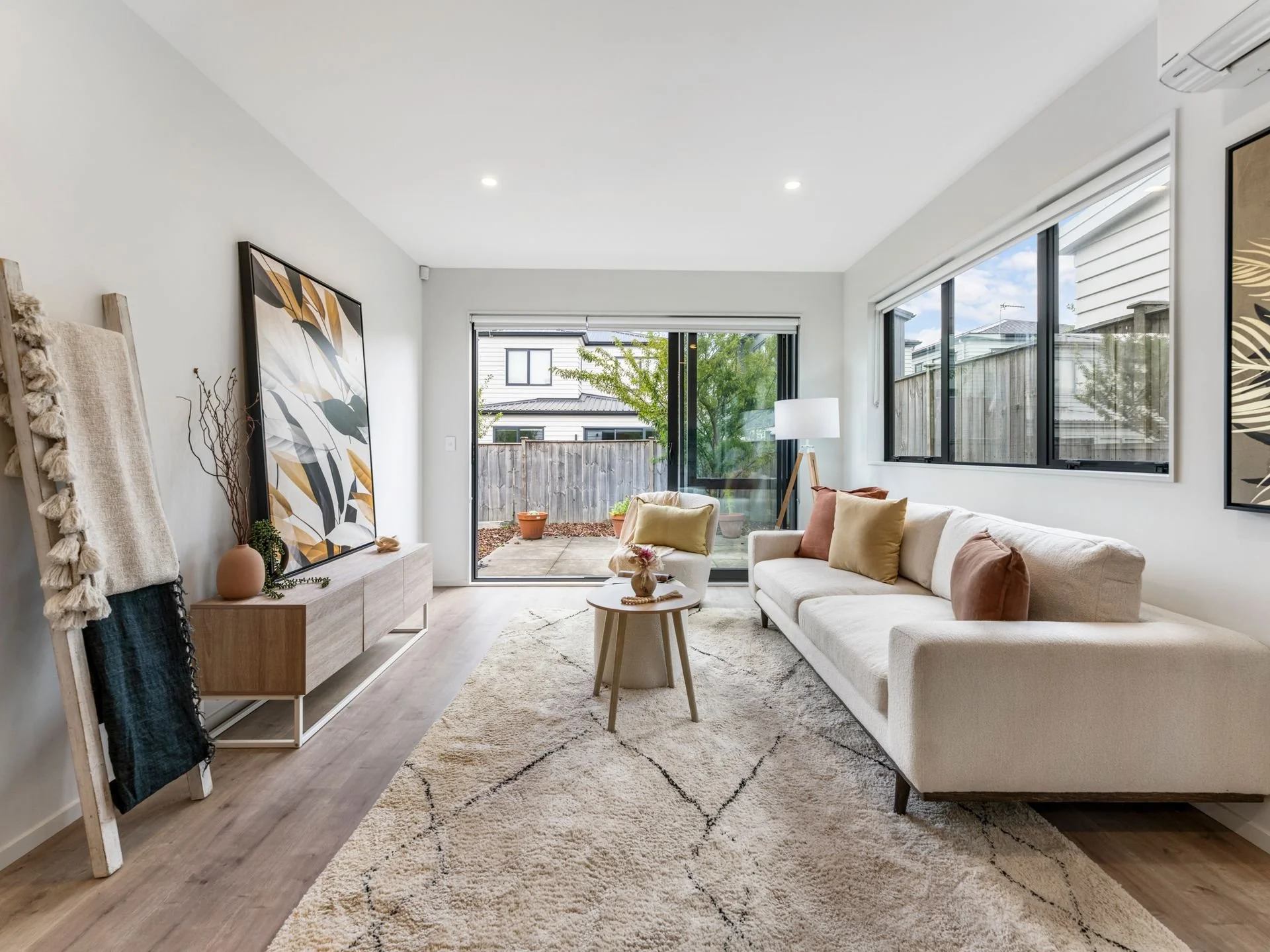 Bright living room with off-white sofa, colorful throw pillows, wooden coffee table, art paintings, sliding glass doors leading to backyard, large windows, and neutral-toned decor.