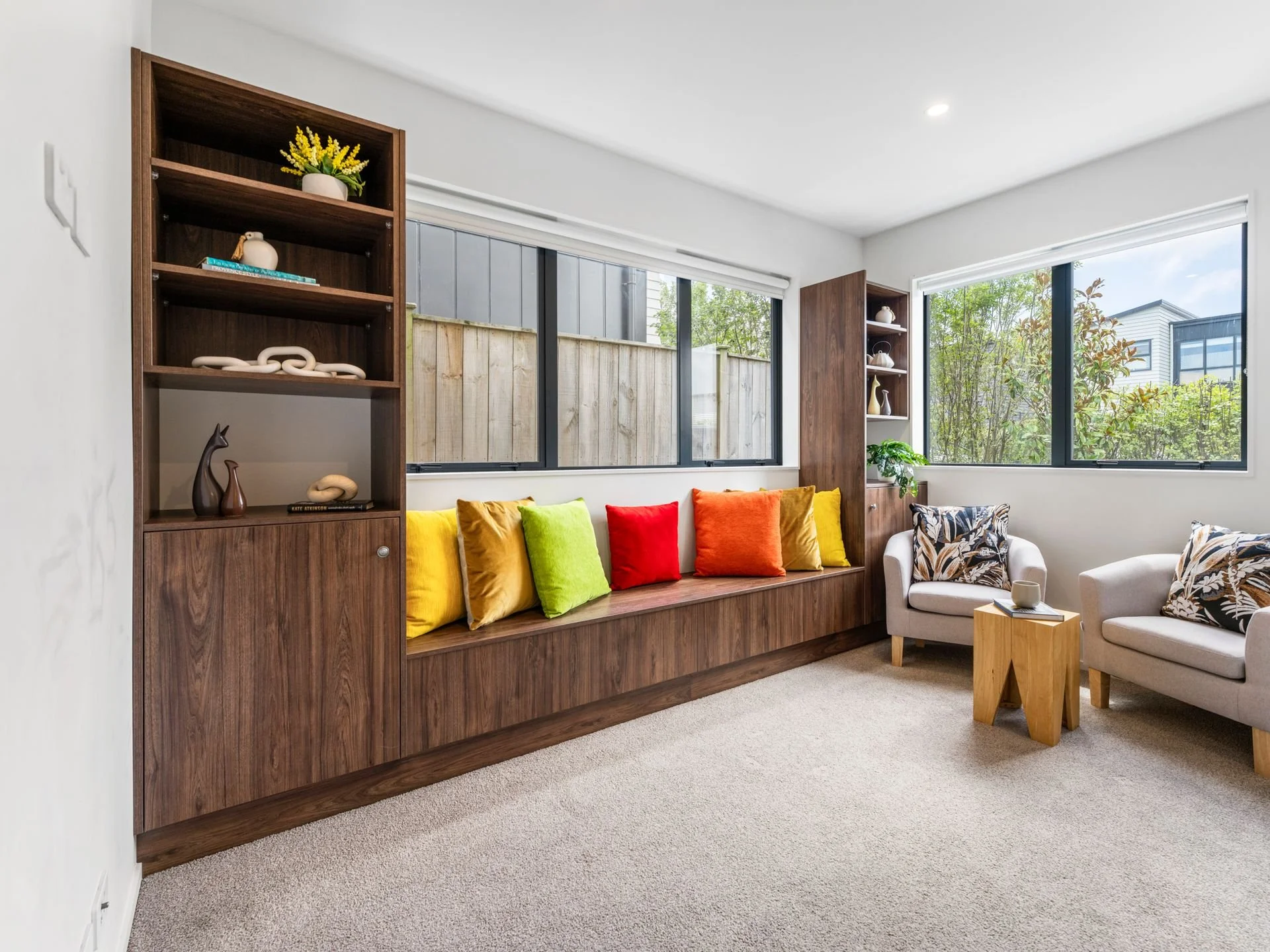 Living room with white walls, beige carpet, two beige armchairs, a small wooden side table, a built-in wooden bench with colorful pillows, large windows showing outdoor greenery, and decorative shelves with plants and decor.