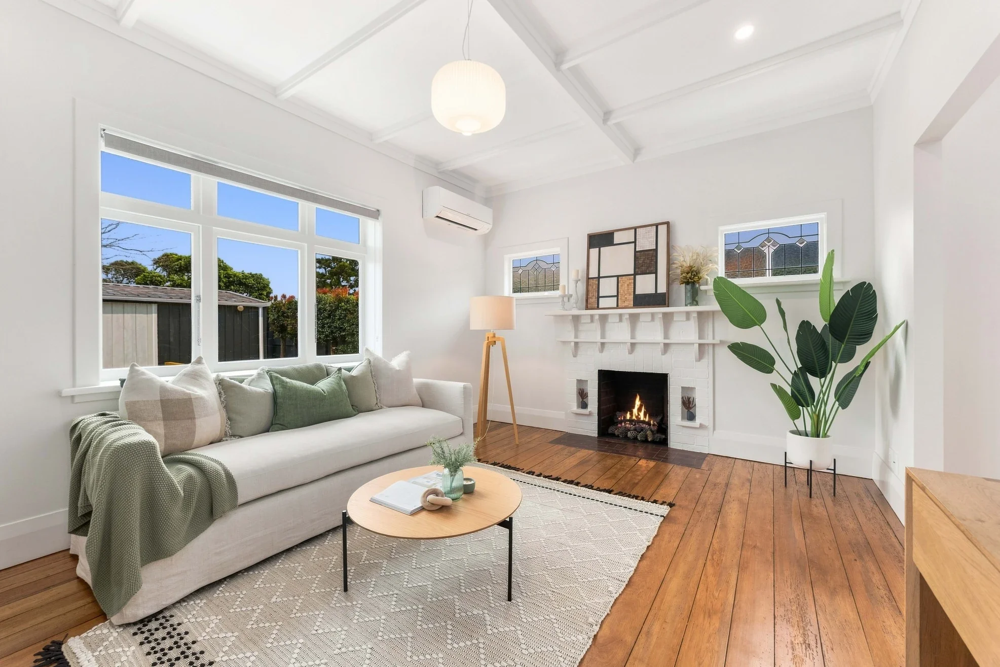Living room with large sliding glass doors showing an outdoor neighborhood sunset, white and beige furniture, a TV stand with artwork, indoor plants, and a carpeted floor.
