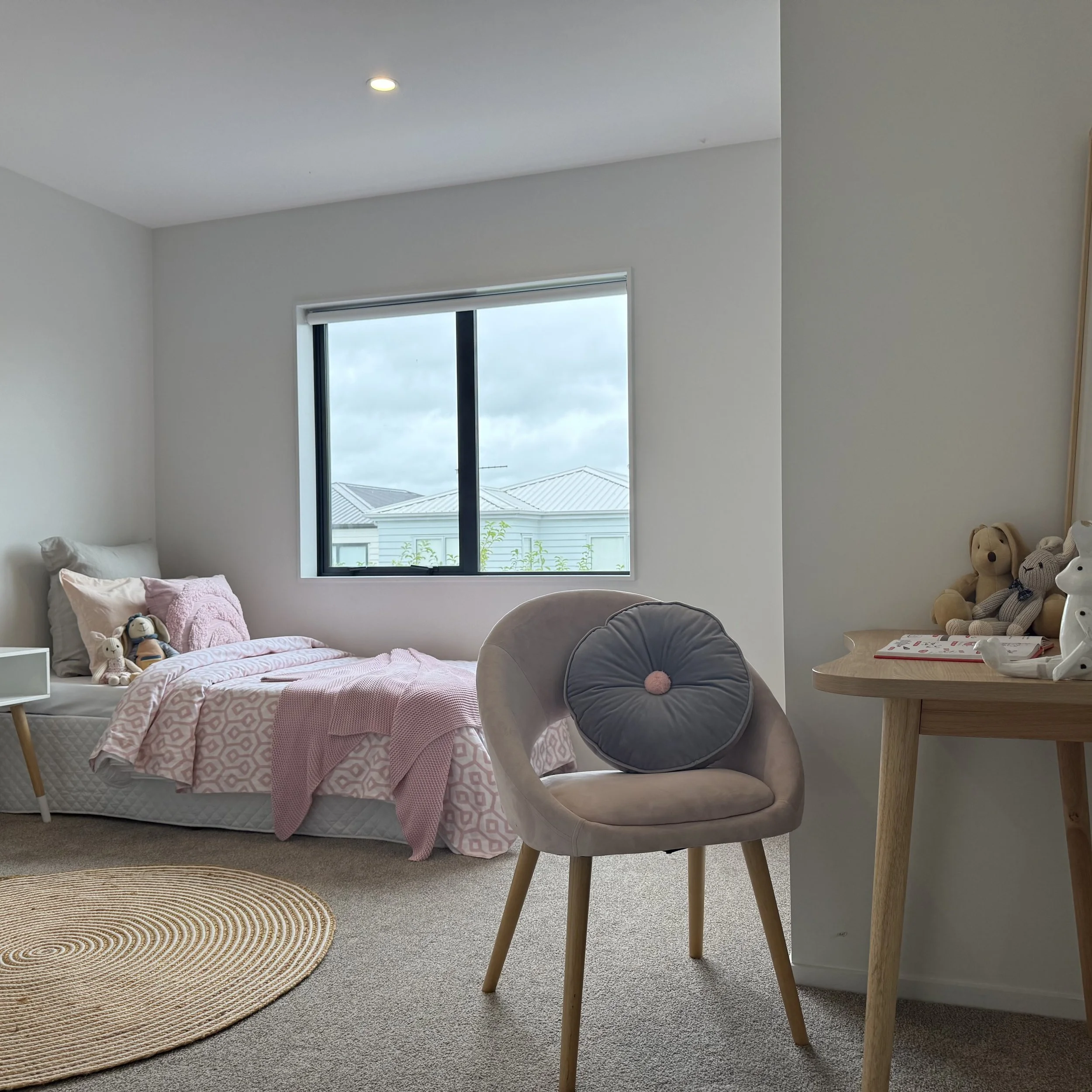 A cozy bedroom with a bed covered in pink and white bedding, stuffed animals on the bed, a beige chair with a round gray pillow, a wooden table with stuffed animals, and a window showing a cloudy sky and rooftops outside.