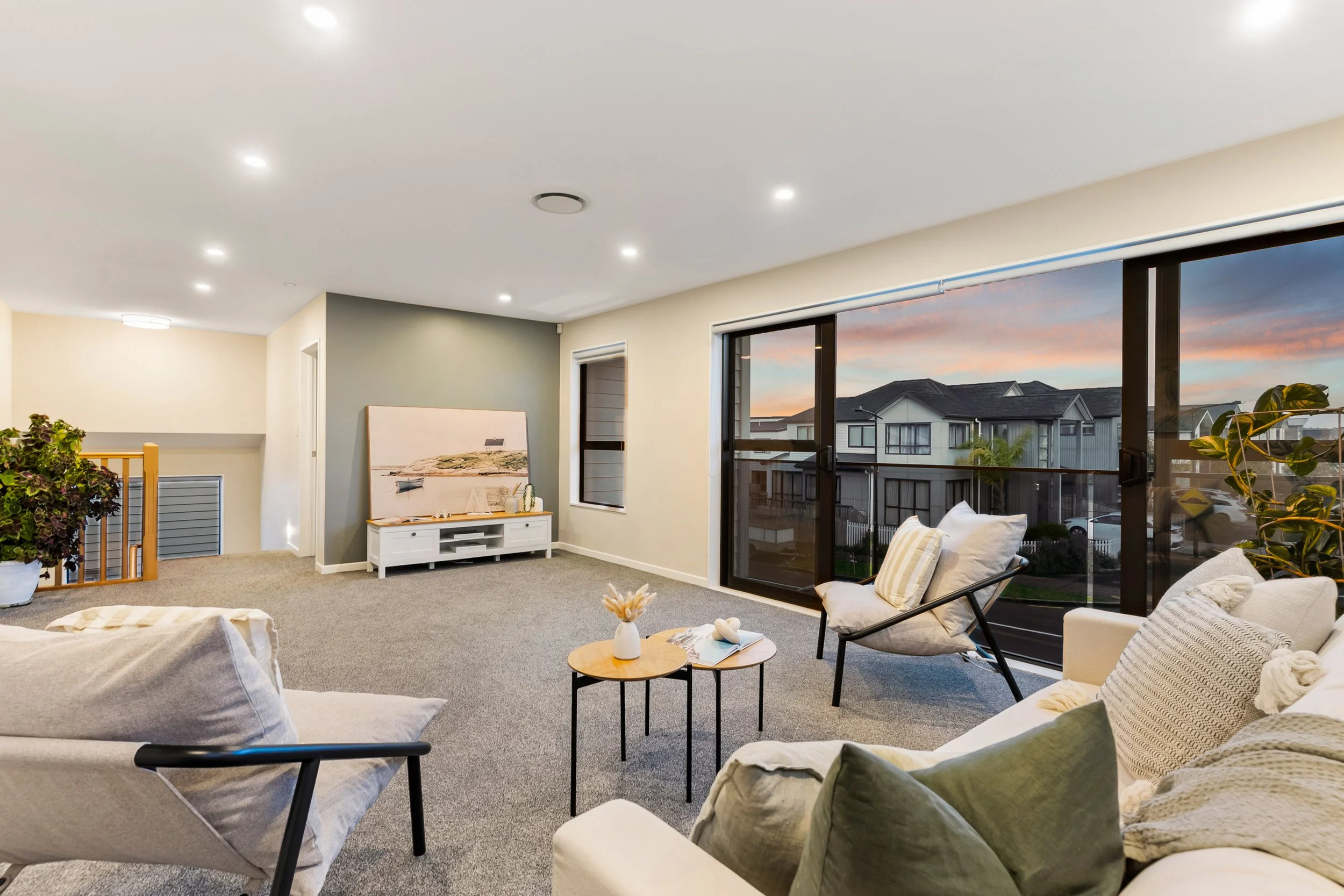 Living room with large sliding glass doors showing an outdoor neighborhood sunset, white and beige furniture, a TV stand with artwork, indoor plants, and a carpeted floor.