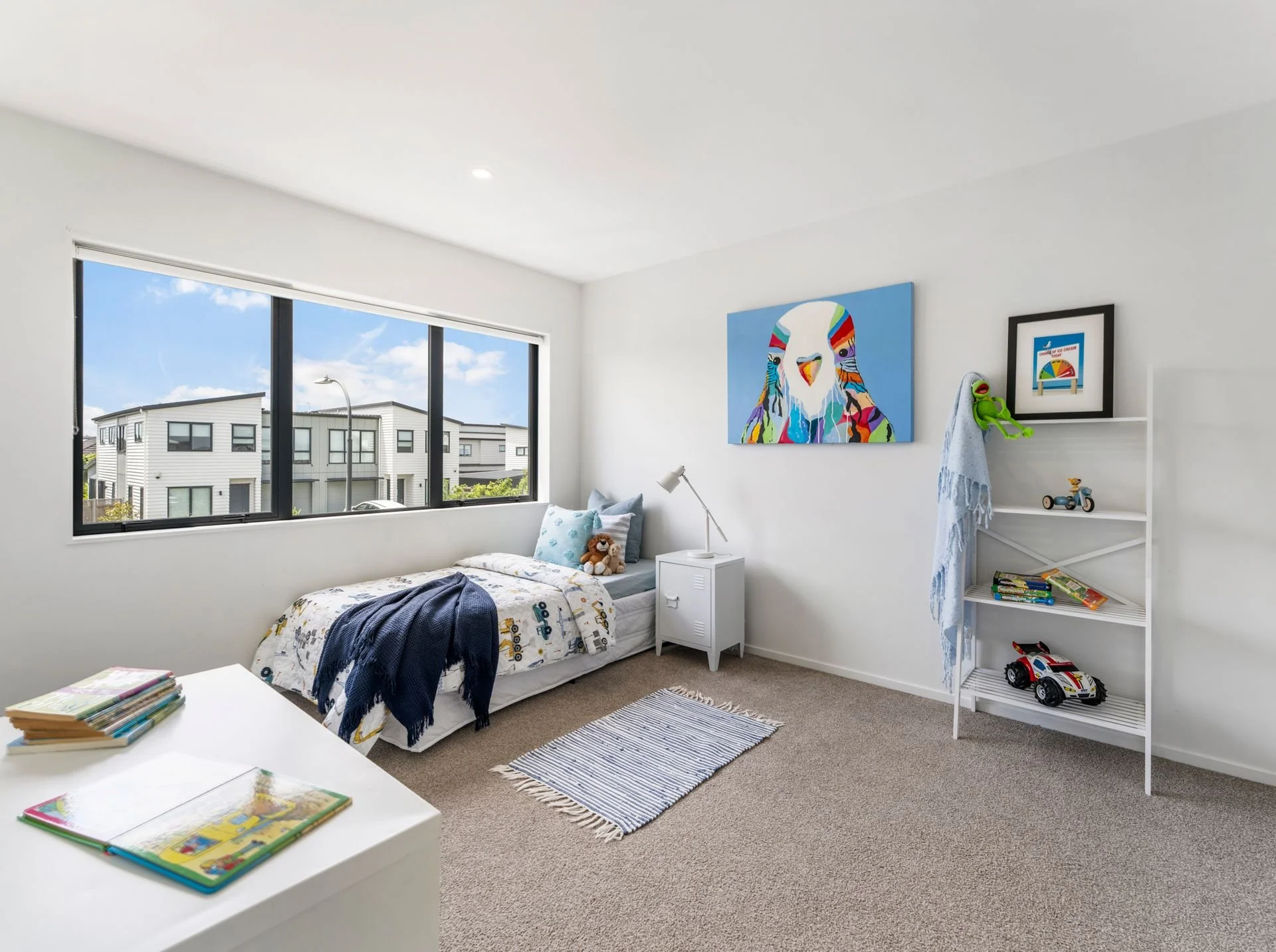 A bright child's bedroom with a single bed covered with a white bedspread featuring colorful vehicles, placed against a large window showing a neighborhood of modern white houses. A dark blue blanket is draped over the bed. A white nightstand with a 