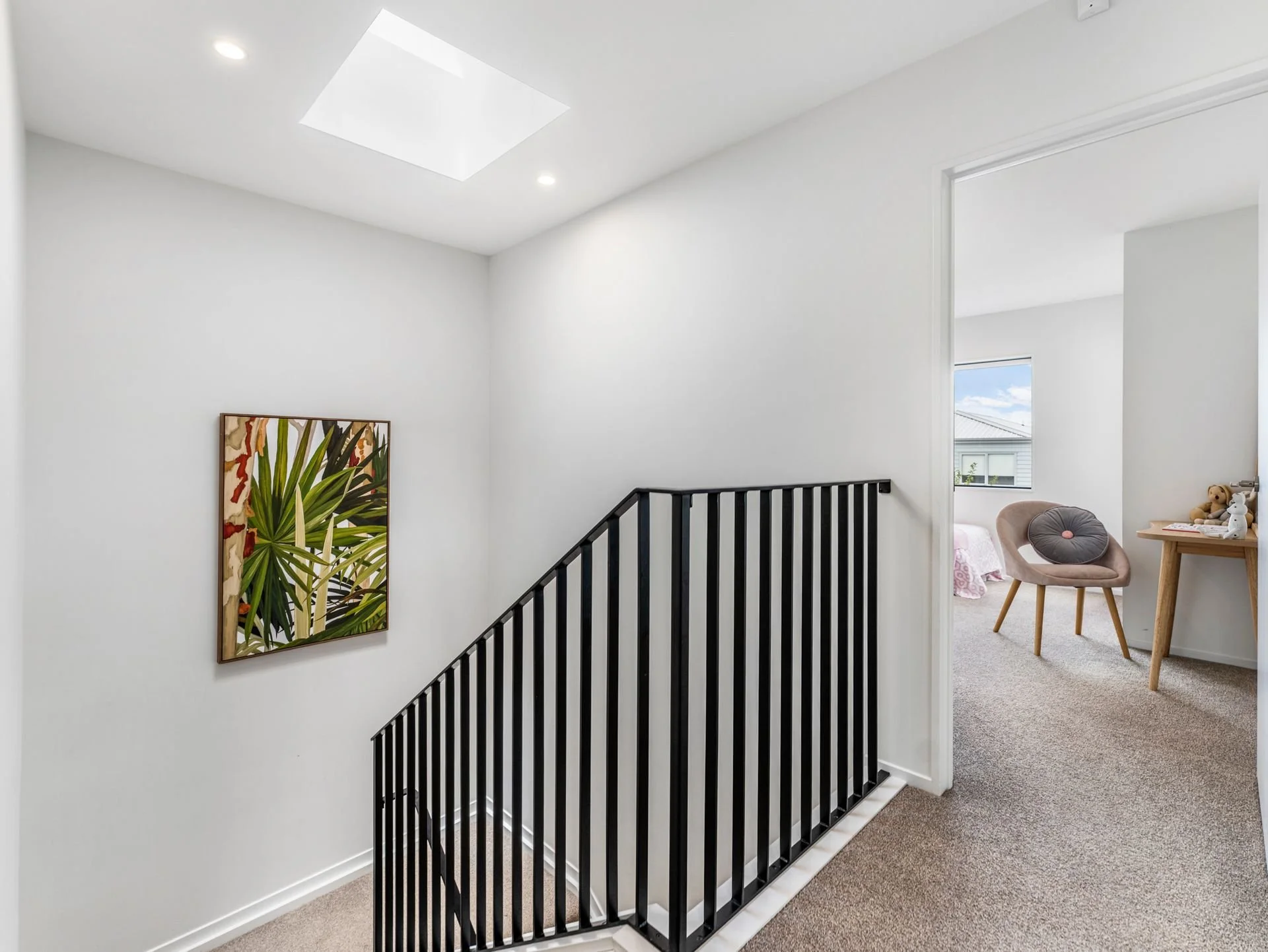 A bright, modern upstairs hallway with white walls, a black metal railing, a ceiling light, a framed botanical artwork, and a view into a bedroom with a chair, a window, and a small desk.