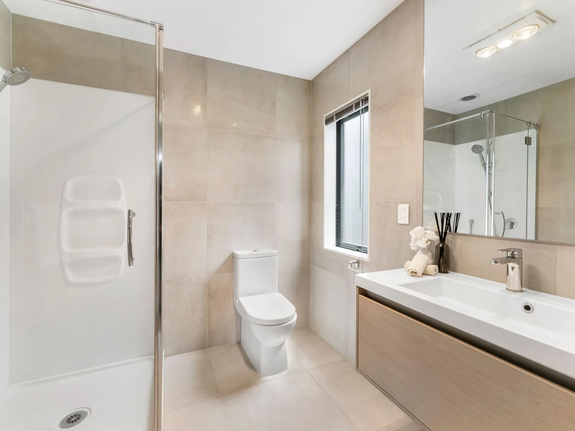 Modern bathroom with a shower stall, toilet, and sink. Beige tiles on walls and floor, window with blinds, large mirror above sink, and minimalist decor with toiletries and a reed diffuser.