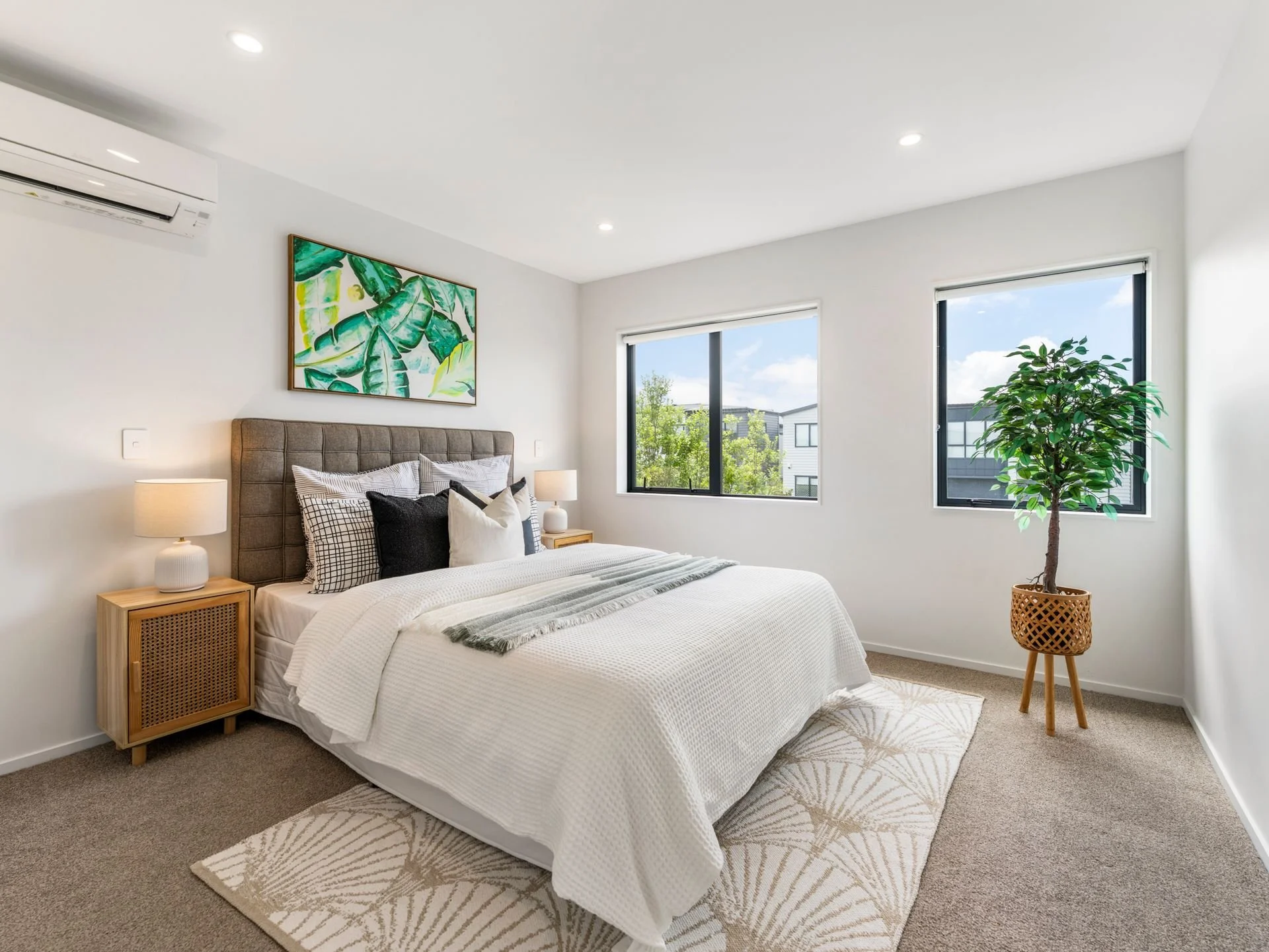 A modern bedroom with white walls, a bed with white bedding and black and white pillows, a wooden headboard, two nightstands with lamps, a potted plant, windows with view of neighboring buildings, a colorful leaf painting, and a beige rug.