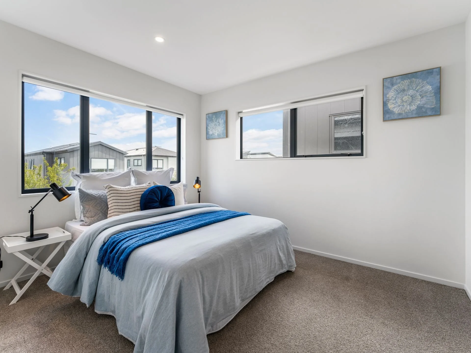 Bedroom with large window overlooking houses and blue sky, bed with white and blue bedding, black bedside lamps, white bedside table, and framed artwork of shells and coral on white walls.