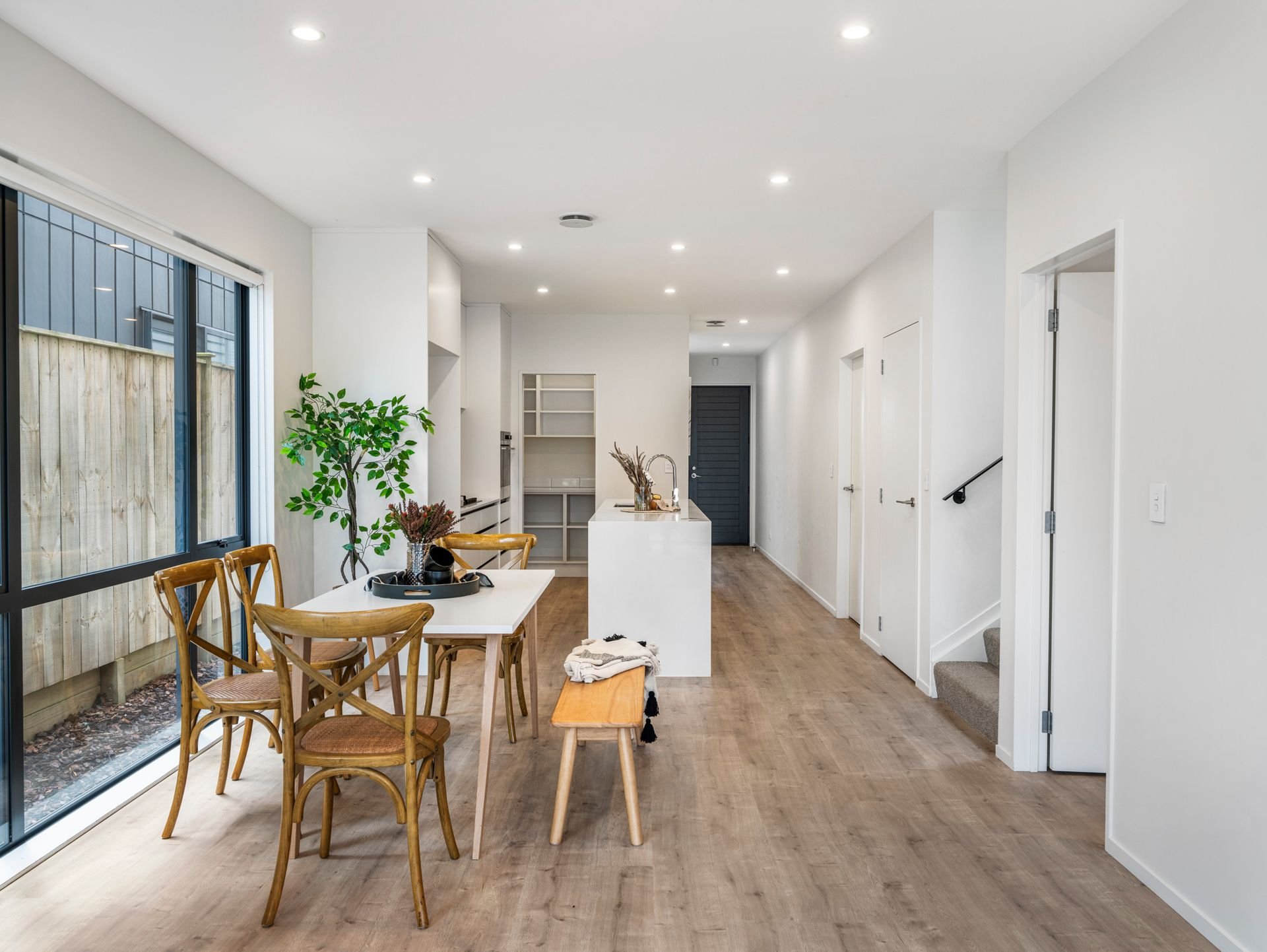 Open-concept kitchen and dining area with wooden flooring, white walls, a large window, a white dining table with wooden chairs, a green potted plant, and a white kitchen island.