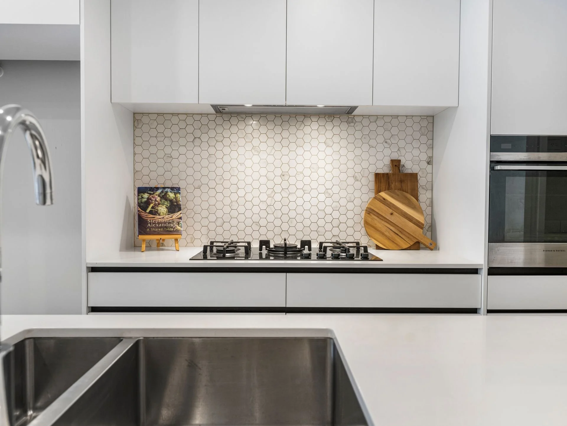 Modern kitchen with white cabinets, hexagonal tile backsplash, and a built-in stove. On the counter are a cookbook on a small easel and a wooden cutting board with a decorative round wooden piece.