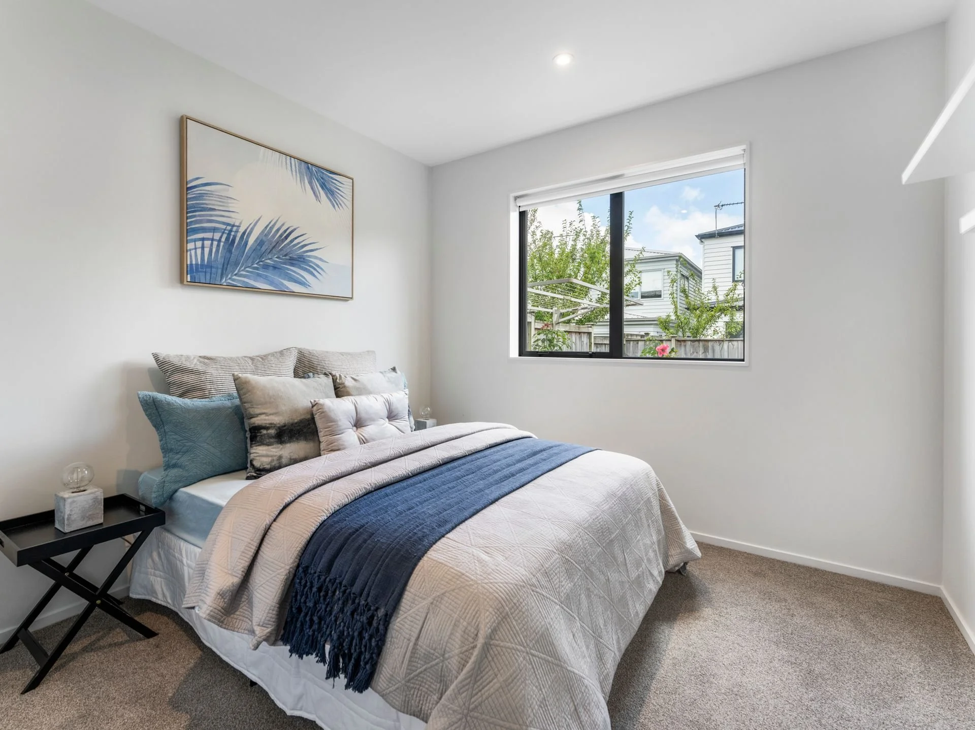 A bedroom with a bed covered in beige and blue linens, multiple pillows, a black nightstand with a glass lamp, a window showing trees and neighboring houses, and a framed rainforest print on the wall.