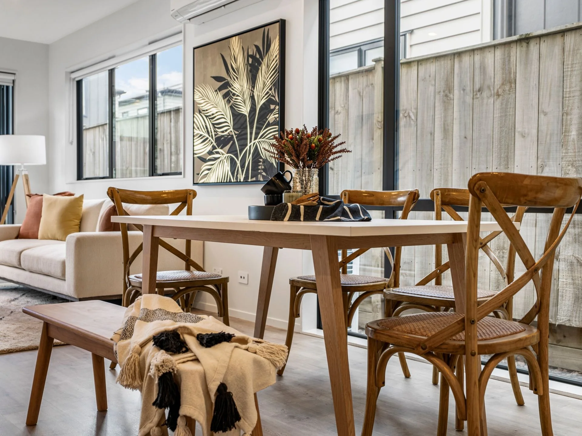 Modern dining area with wooden table and chairs, a beige sofa with pillows, a large botanical art piece on the wall, and large windows.