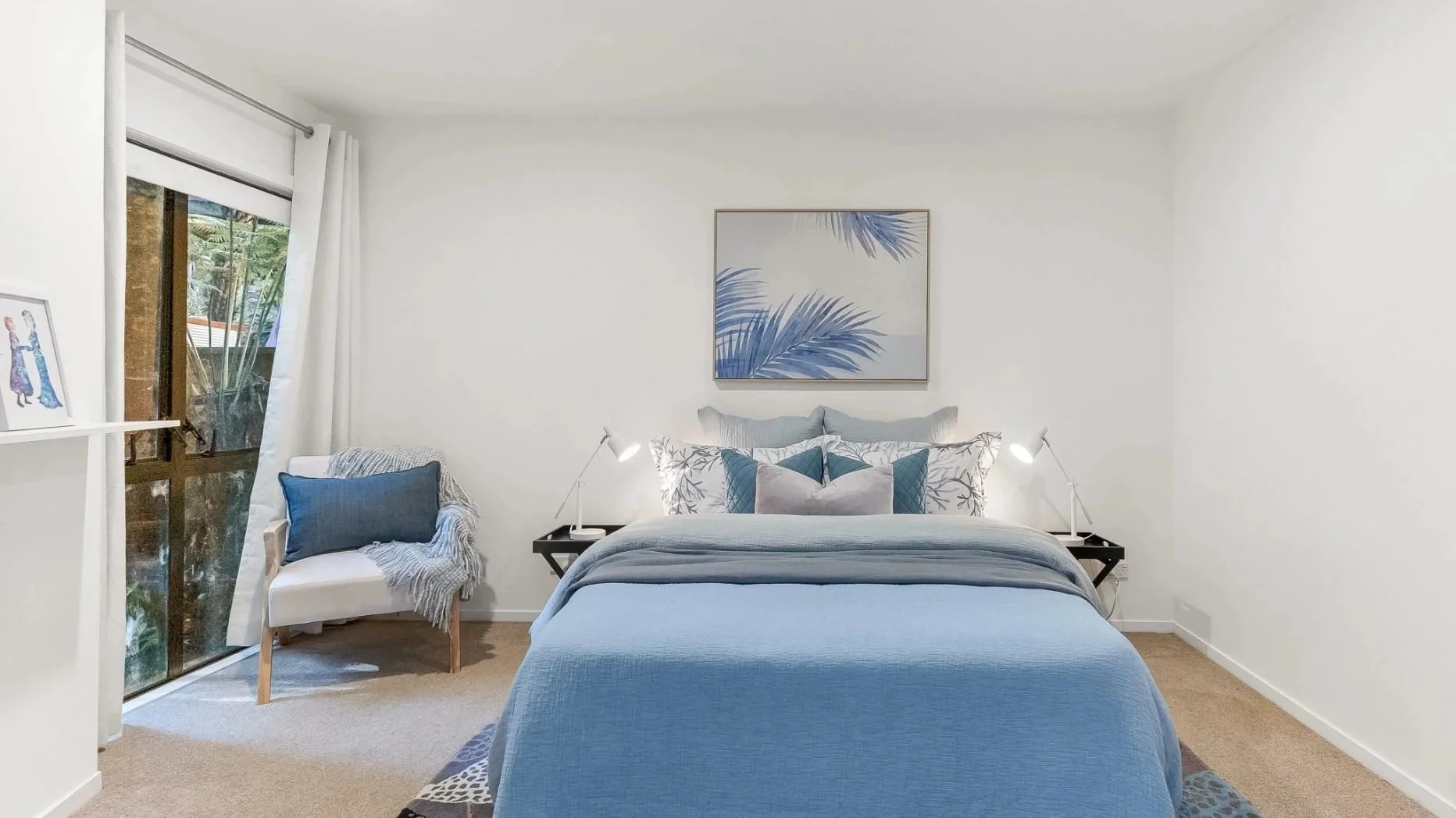 A modern bedroom with white walls and beige carpet, featuring a bed with blue bedding and matching pillows, a picture of leaves on the wall, two white bedside lamps, a white chair with a blue pillow and blanket, and a sliding glass door with white curtains leading outside.