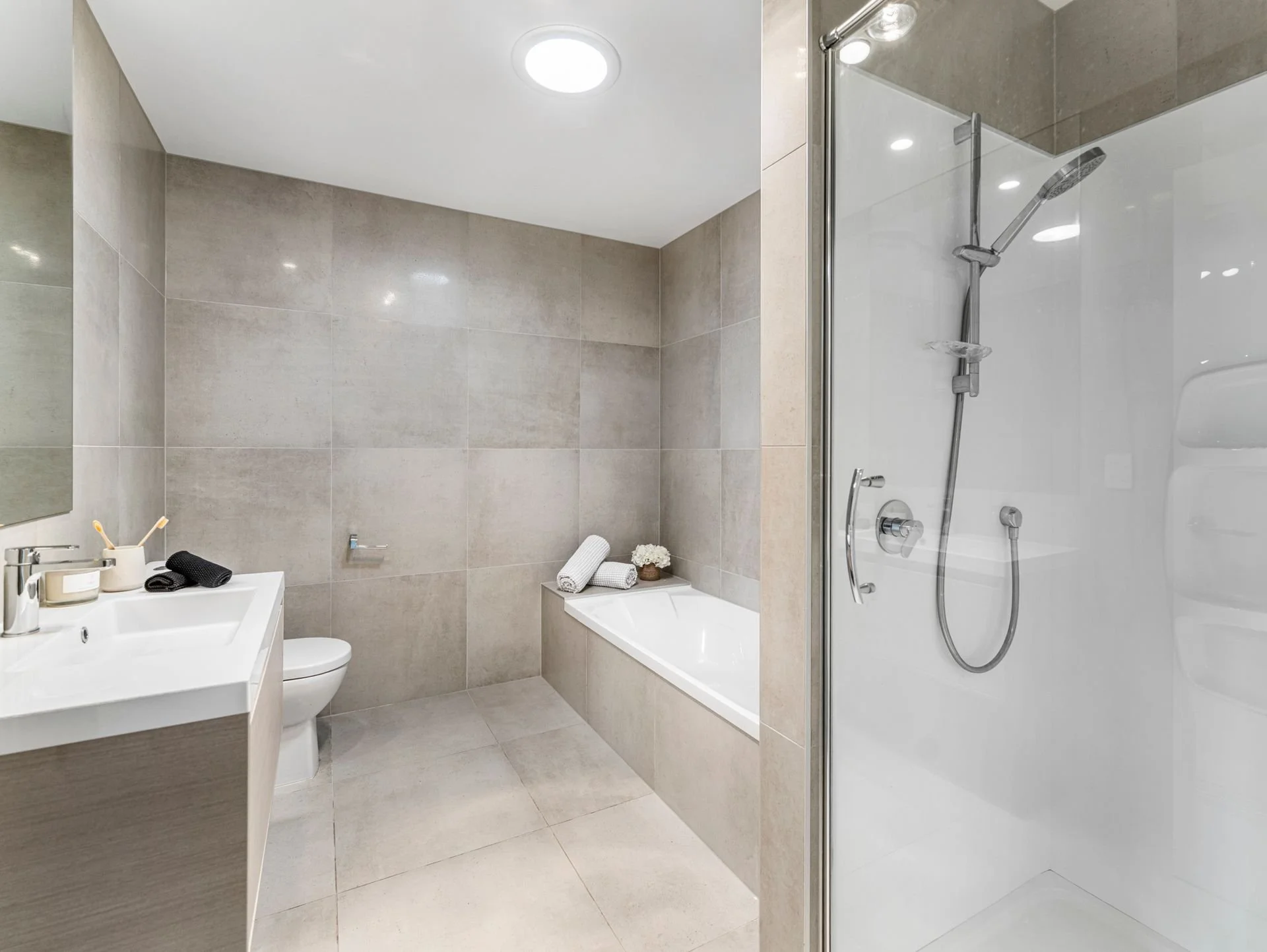 Modern bathroom with beige tiled walls and floor, a white bathtub with towels and flowers on its edge, a white sink with toiletries, a toilet, and a glass-enclosed shower with a handheld showerhead.