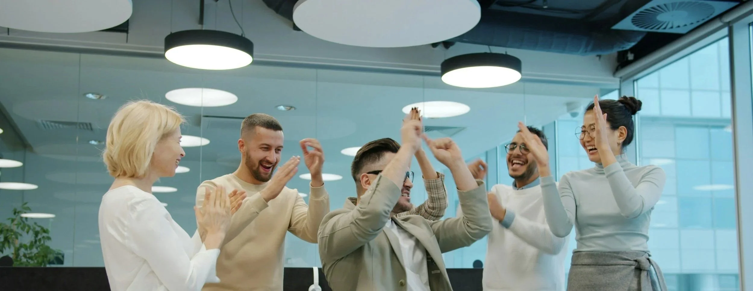 Group of five diverse colleagues celebrating with high-fives in an office, smiling and dressed in business casual attire.