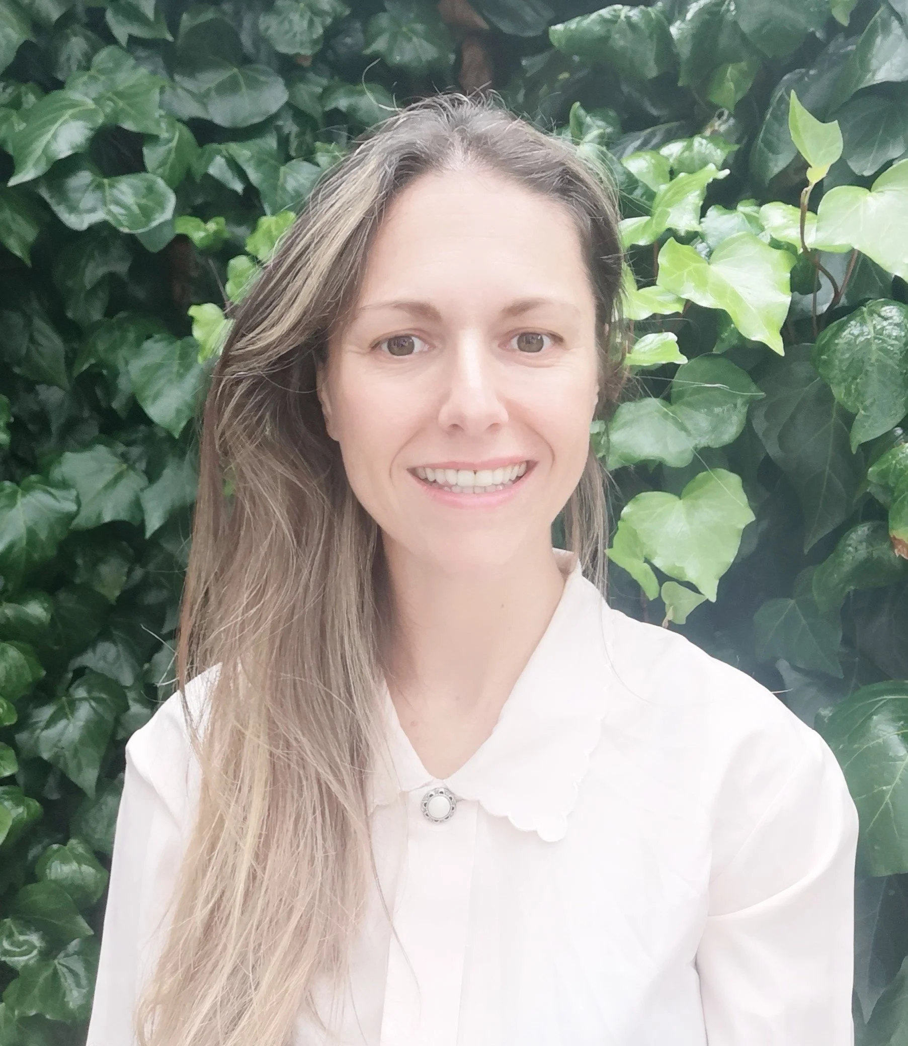 Close-up portrait of a smiling woman with long wavy hair, wearing a white collared shirt, standing in front of a lush green leafy background.