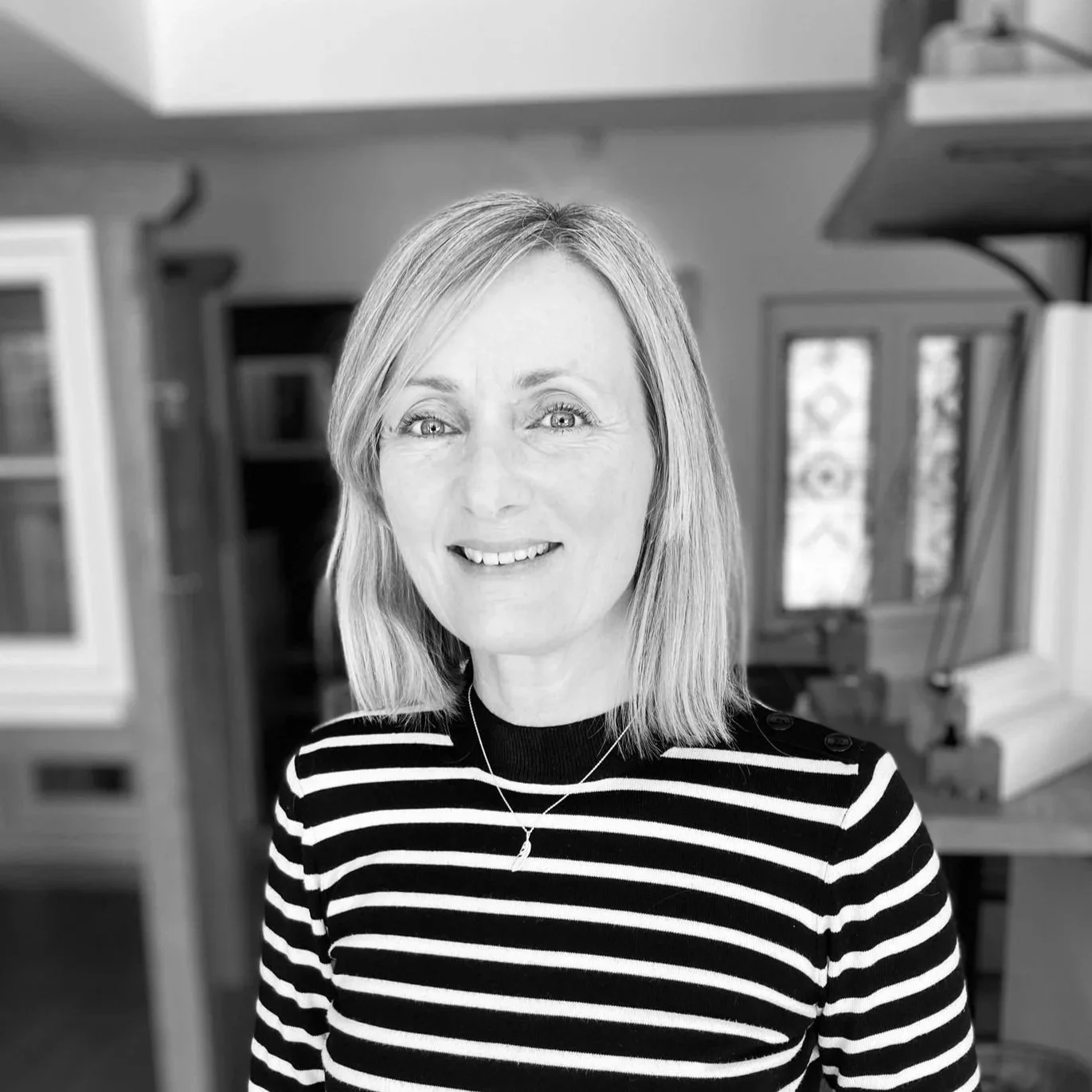 Black and white portrait of a woman with shoulder-length hair wearing a striped shirt and a necklace, standing in a room with large windows and furniture in the background.