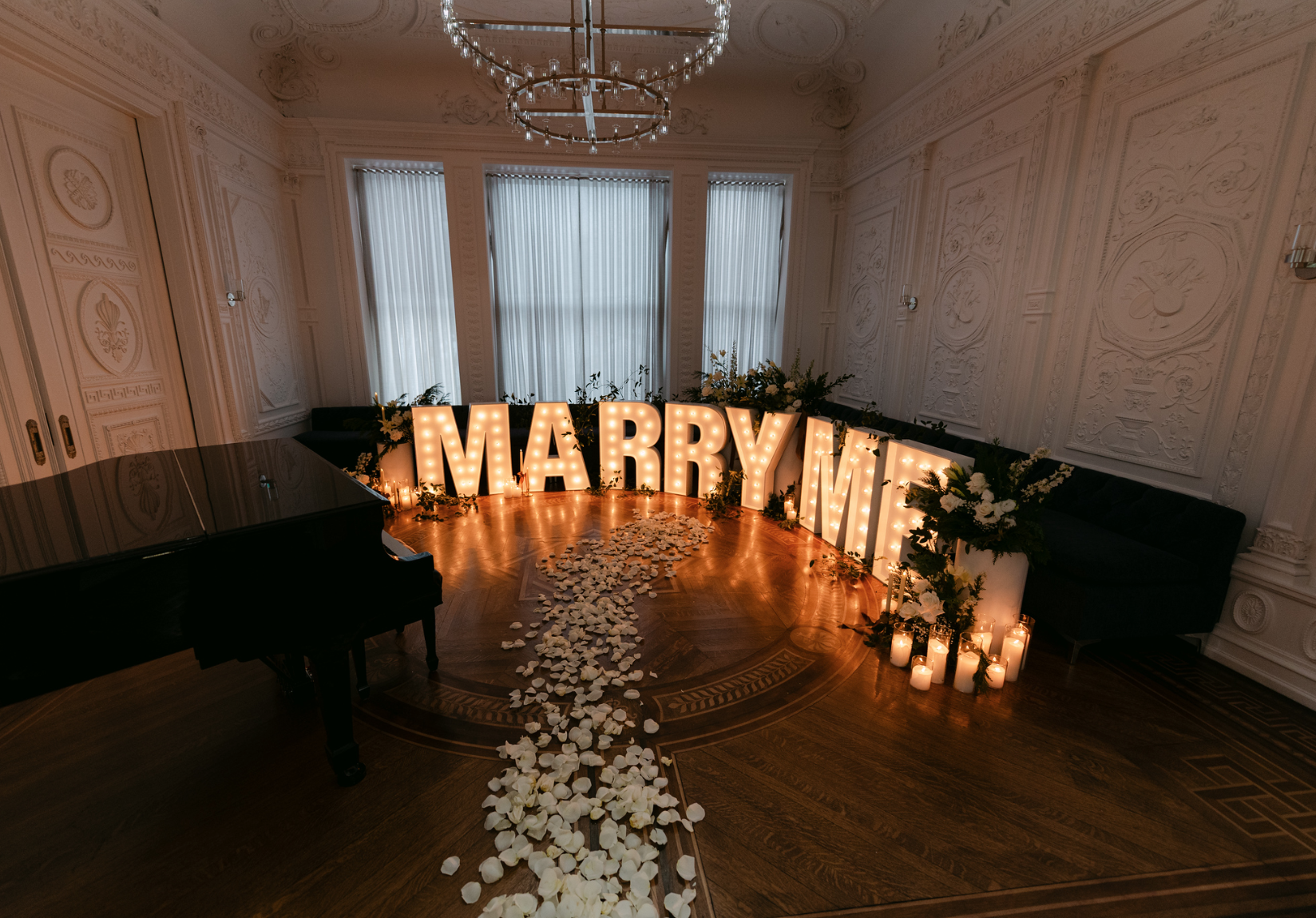Lit letters spelling Marry Me sit on the ground with candles and a path of petals