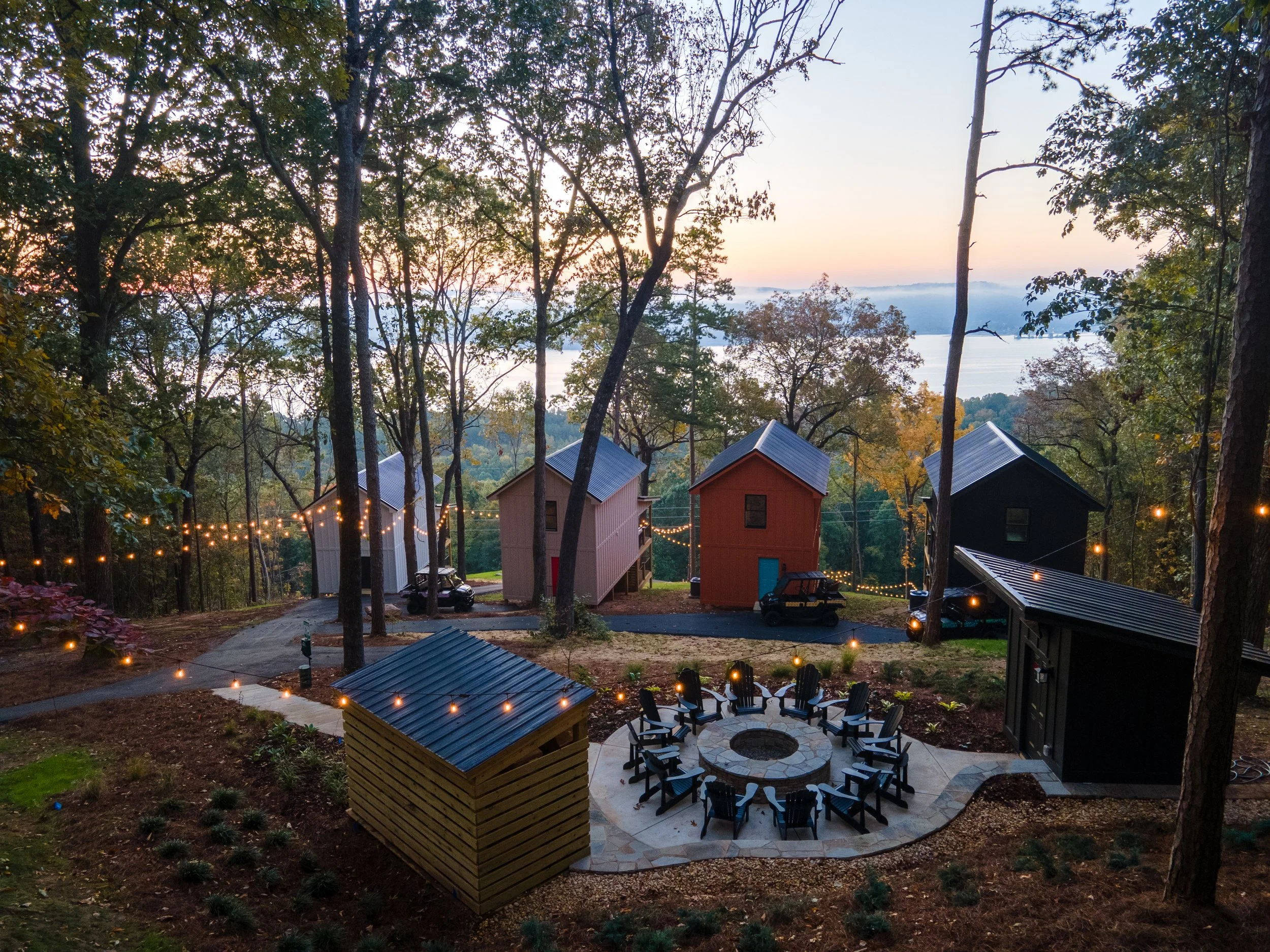 Cabins by the lake at dusk with trees and string lights hanging across the yard around a circular fire pit area with chairs.