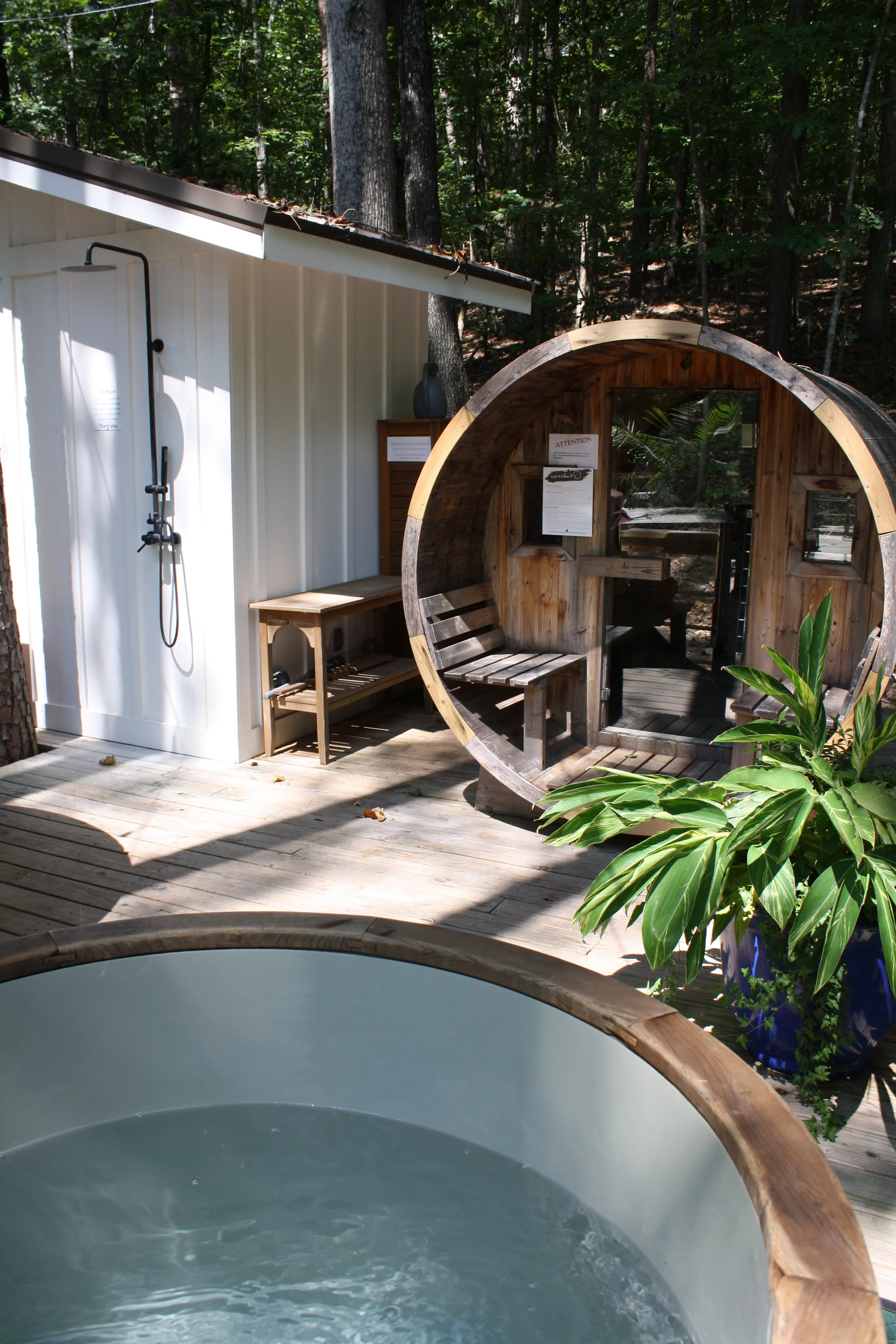 Outdoor hot tub with a wooden rim, surrounded by a deck with a potted plant, an outdoor shower, a wooden bench, and a wooden hexagonal cabin with a forest backdrop.