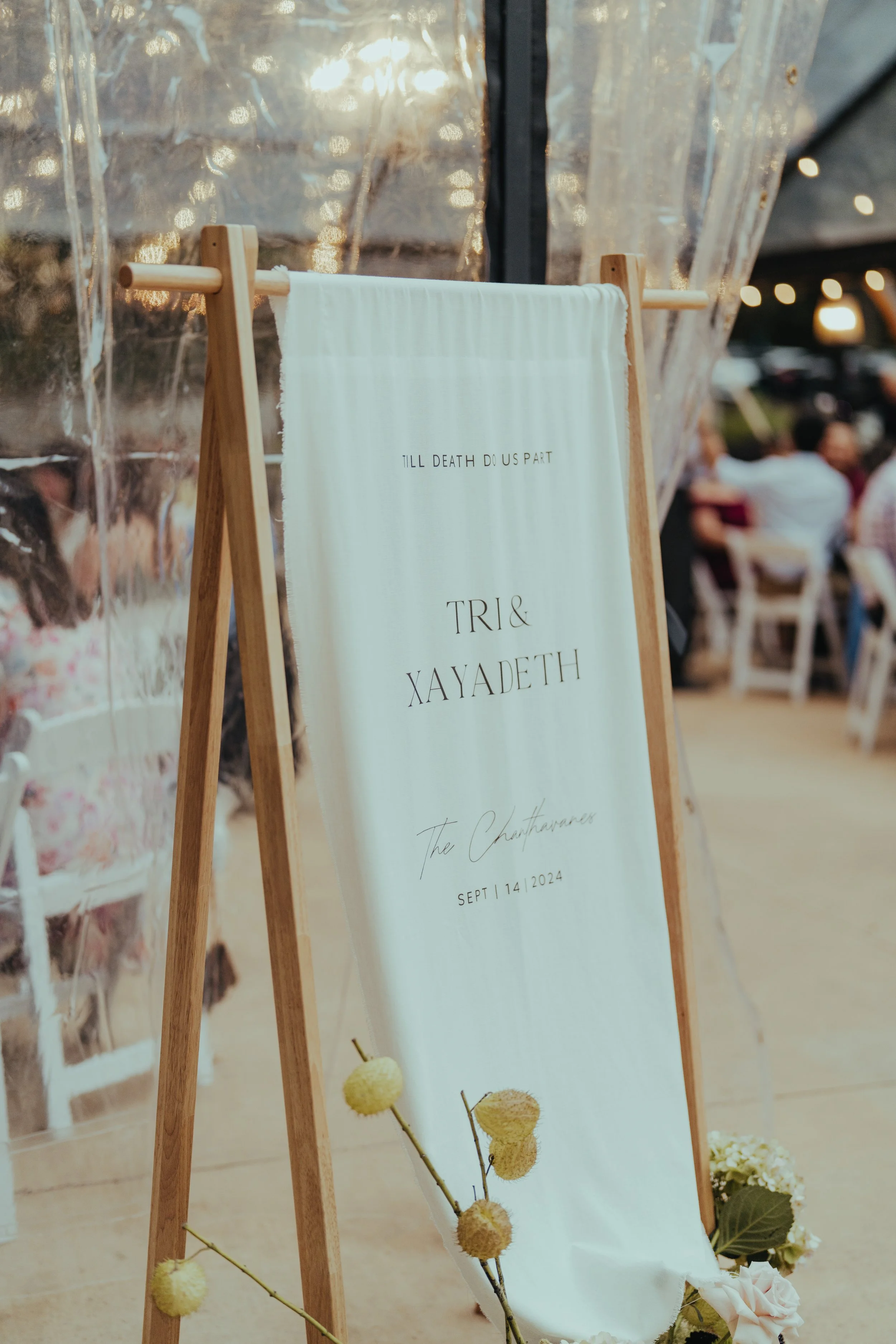 A wedding sign on an easel reads "Till death do us part" and includes the names "Tri & Xayadeth" with the wedding date "September 14, 2024." The background shows blurred guests at an outdoor wedding with sunlight filtering through, and decorative flo