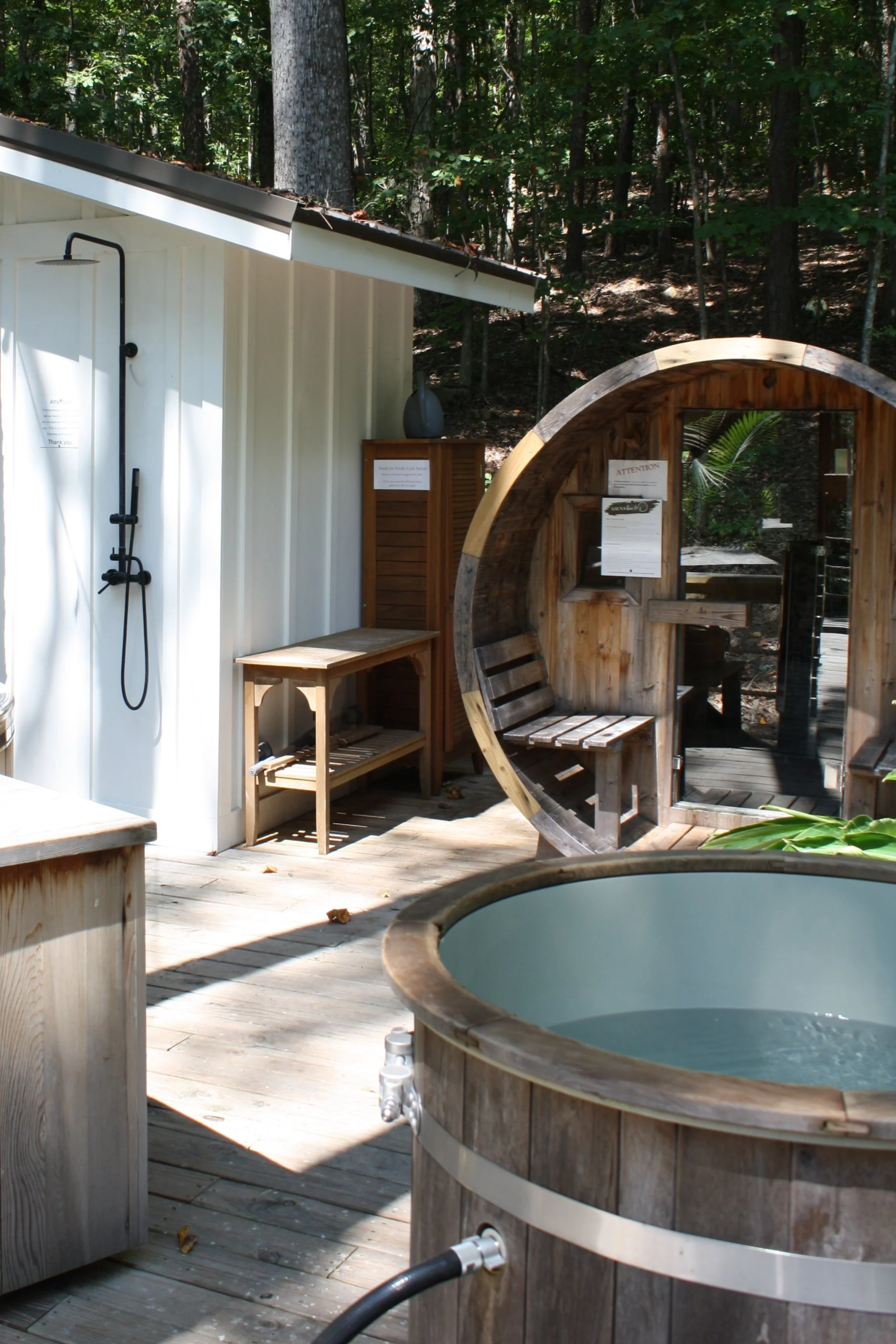 Outdoor wooden shower station and hot tub in a forest setting with trees and sunny weather.