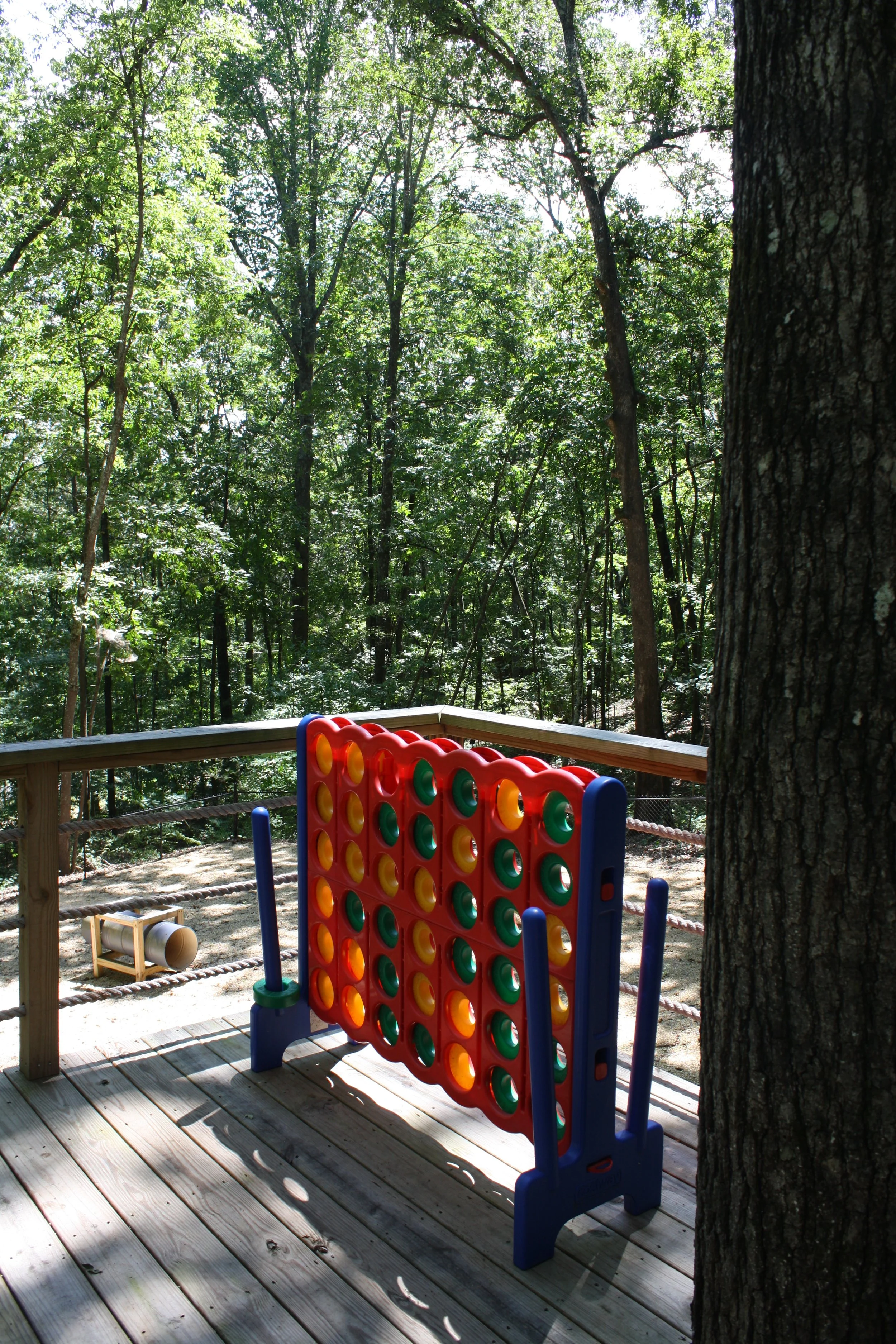 A wooden deck with a large tree trunk on the right and a colorful outdoor Connect 4 game in the foreground. The deck overlooks a lush green forest with tall trees and dense foliage.