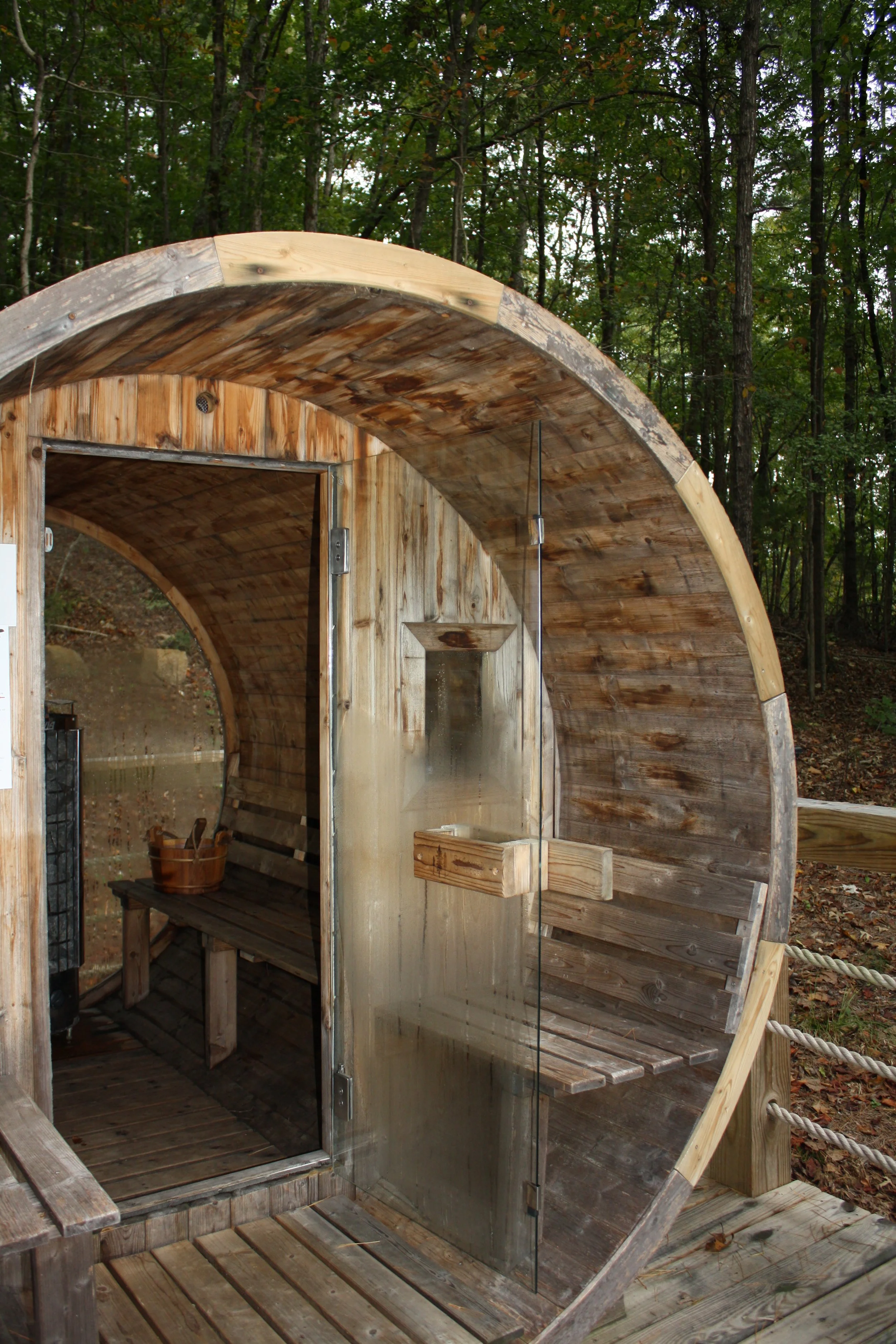 A wooden outdoor sauna with a rounded roof located in a wooded area, with a glass door and interior seating.