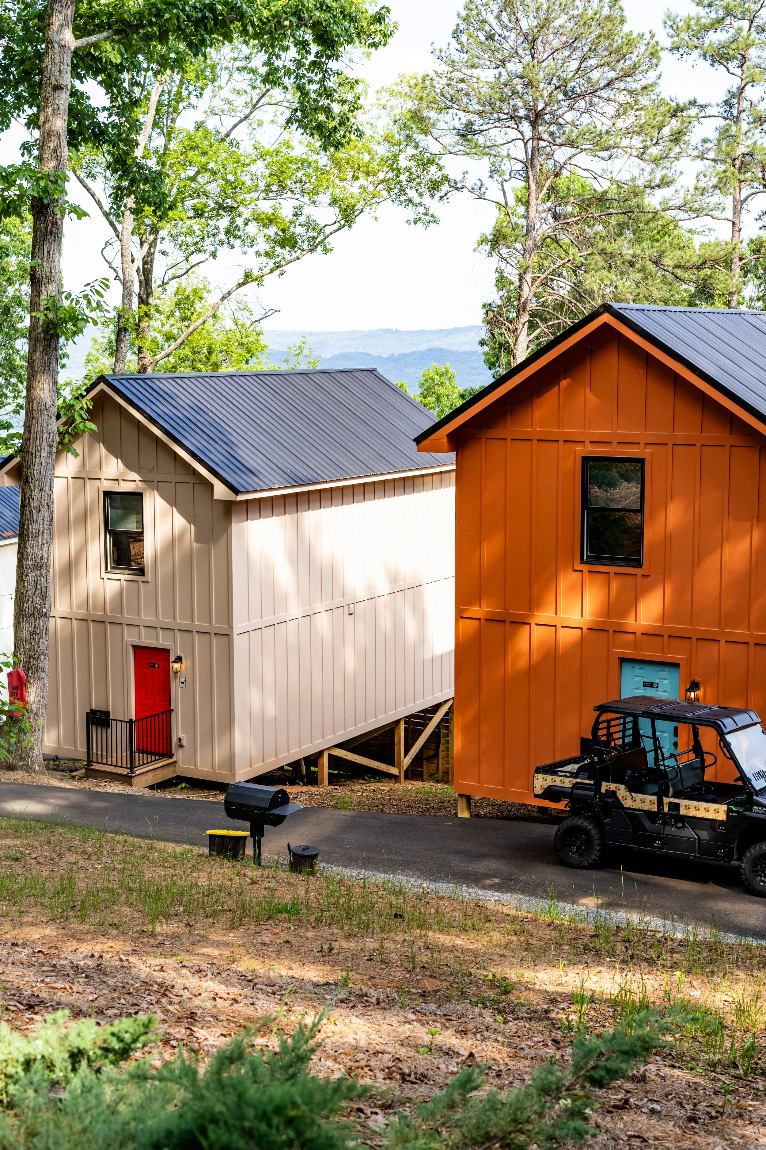 Two colorful cabins, one beige and one orange, surrounded by trees and plants in a lush outdoor setting.