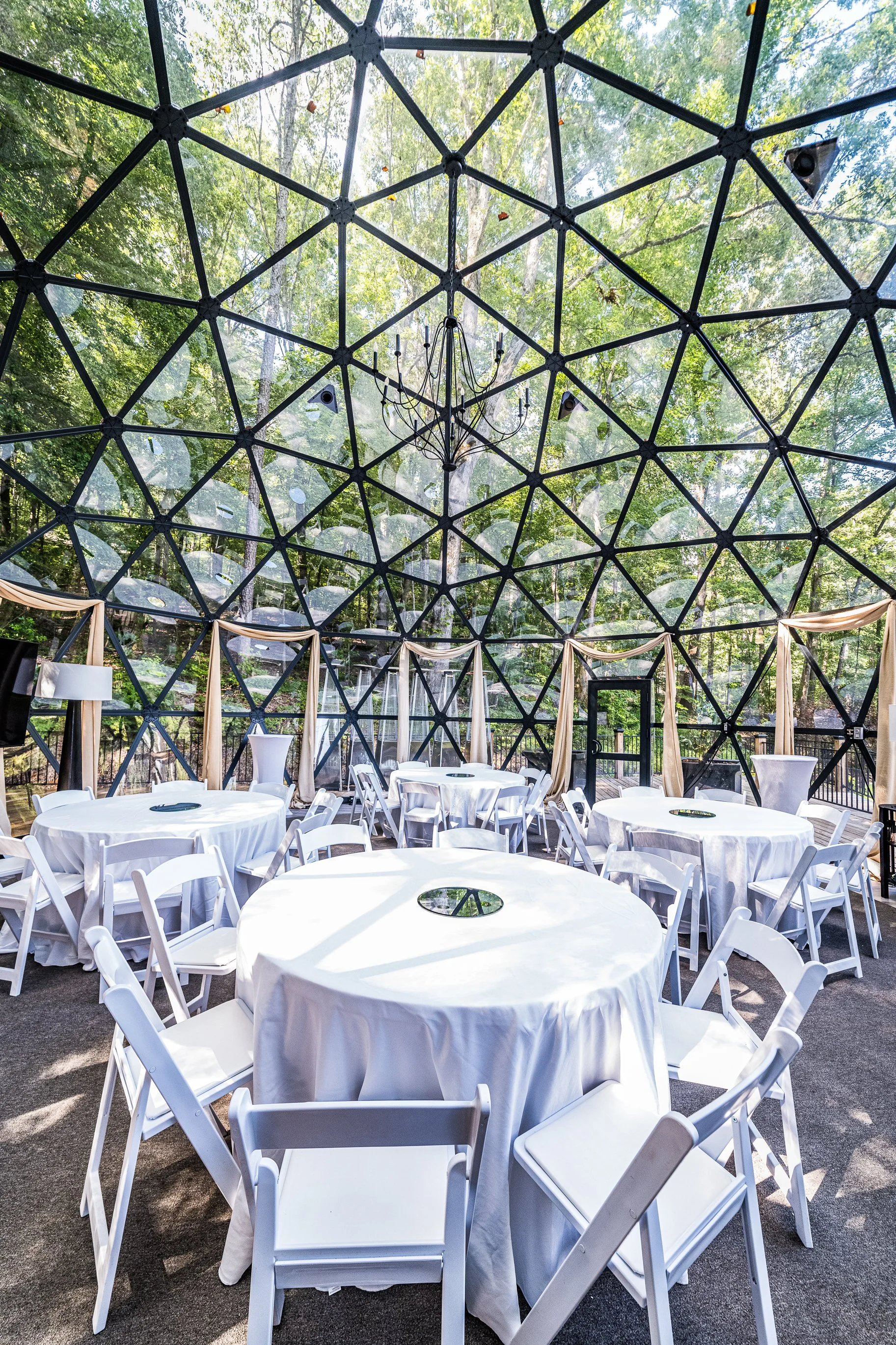 Indoor event space with round tables covered in white tablecloths, surrounded by white chairs, and a glass geodesic dome ceiling showing green trees outside. There are draped beige curtains and a chandelier hanging from the ceiling.