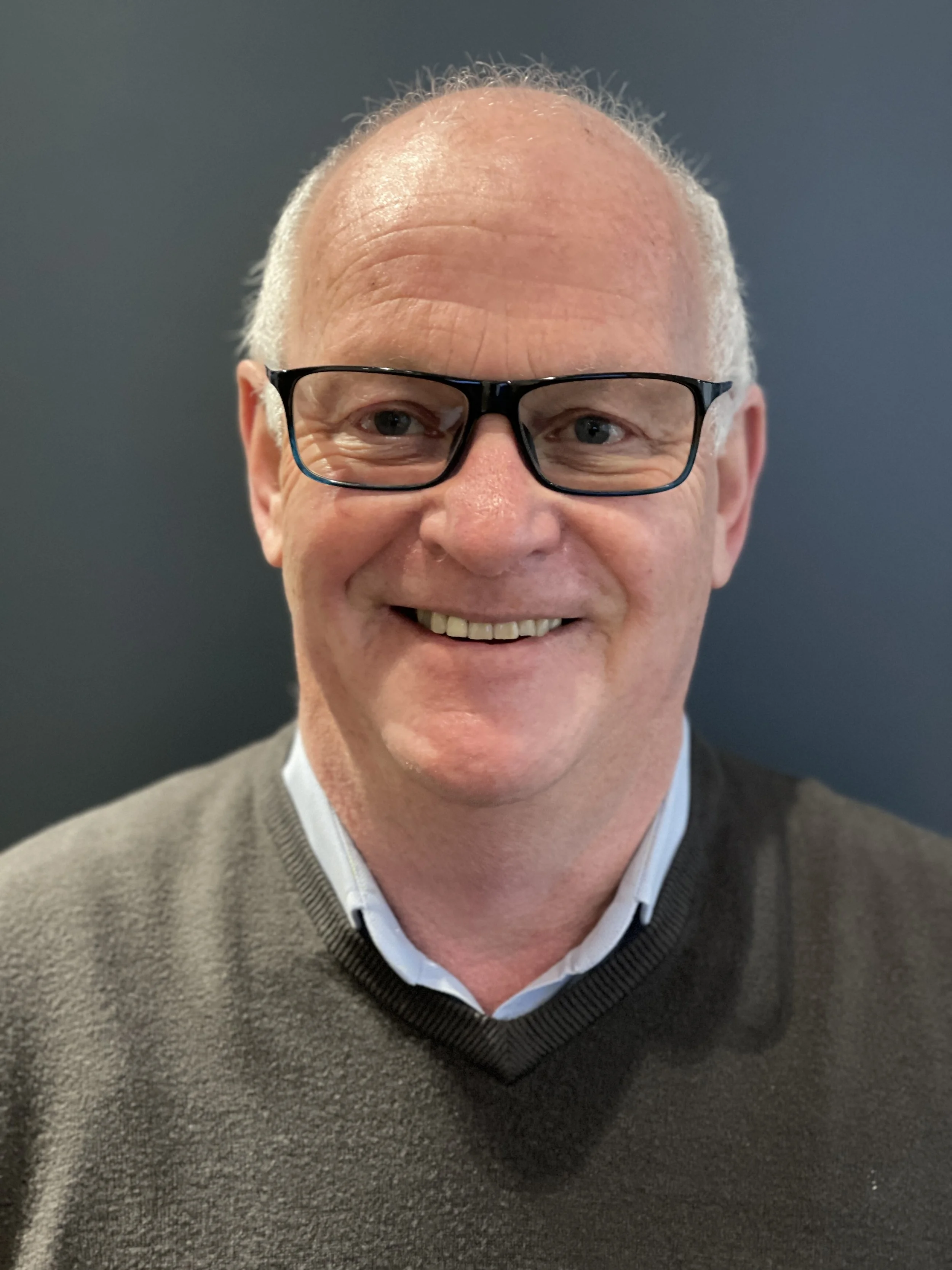 Close-up photo of a smiling middle-aged man with glasses, wearing a dark sweater over a collared shirt, against a dark background.