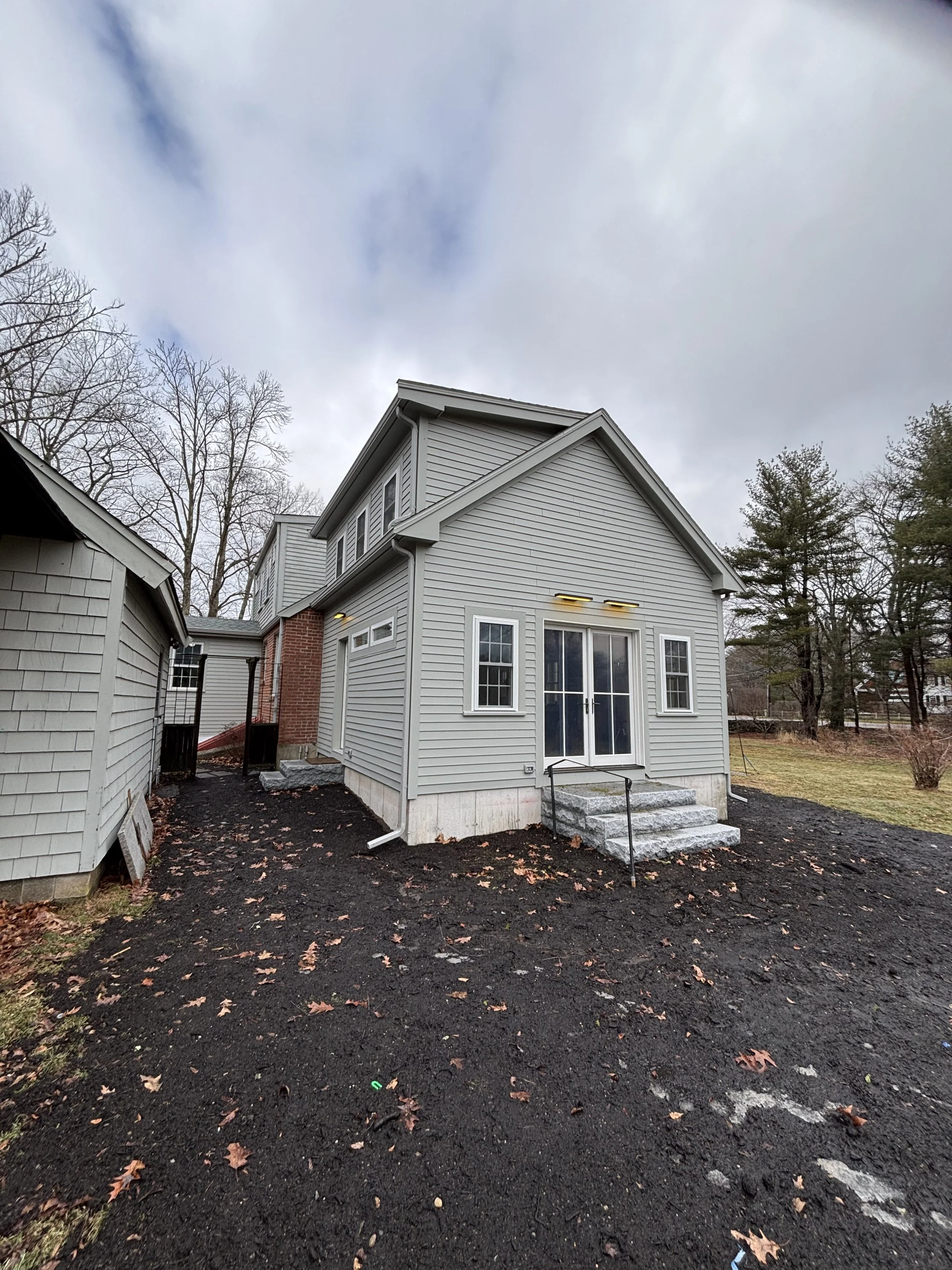 A newly constructed gray house with a small set of concrete steps leading to glass double doors, surrounded by dark soil and sparse fallen leaves, with an overcast sky and leafless trees in the background.