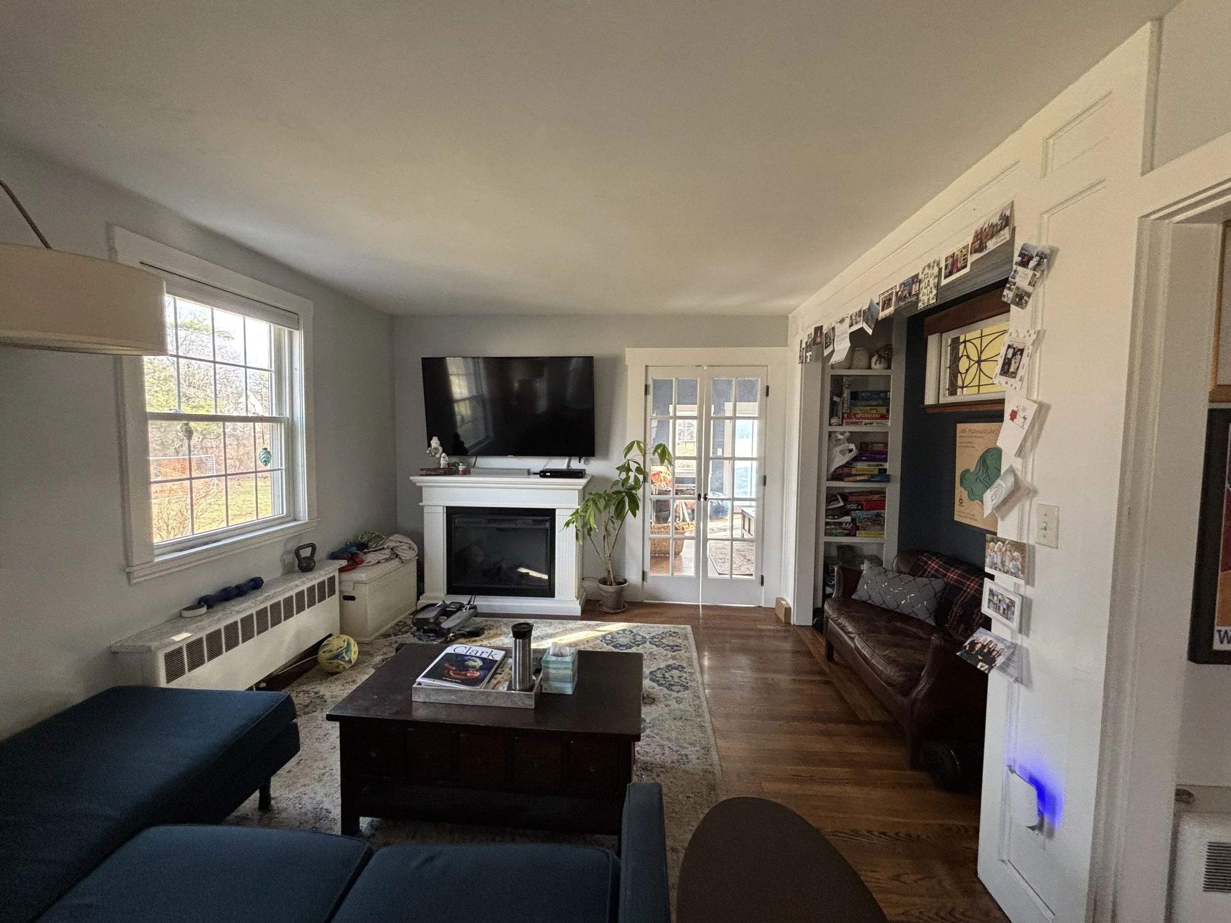 Living room with a large window, a TV mounted above a fireplace, a small daybed, a coffee table with books, and a door leading to an outdoor patio. There is a potted plant near the door and a wall with photos and decorations.