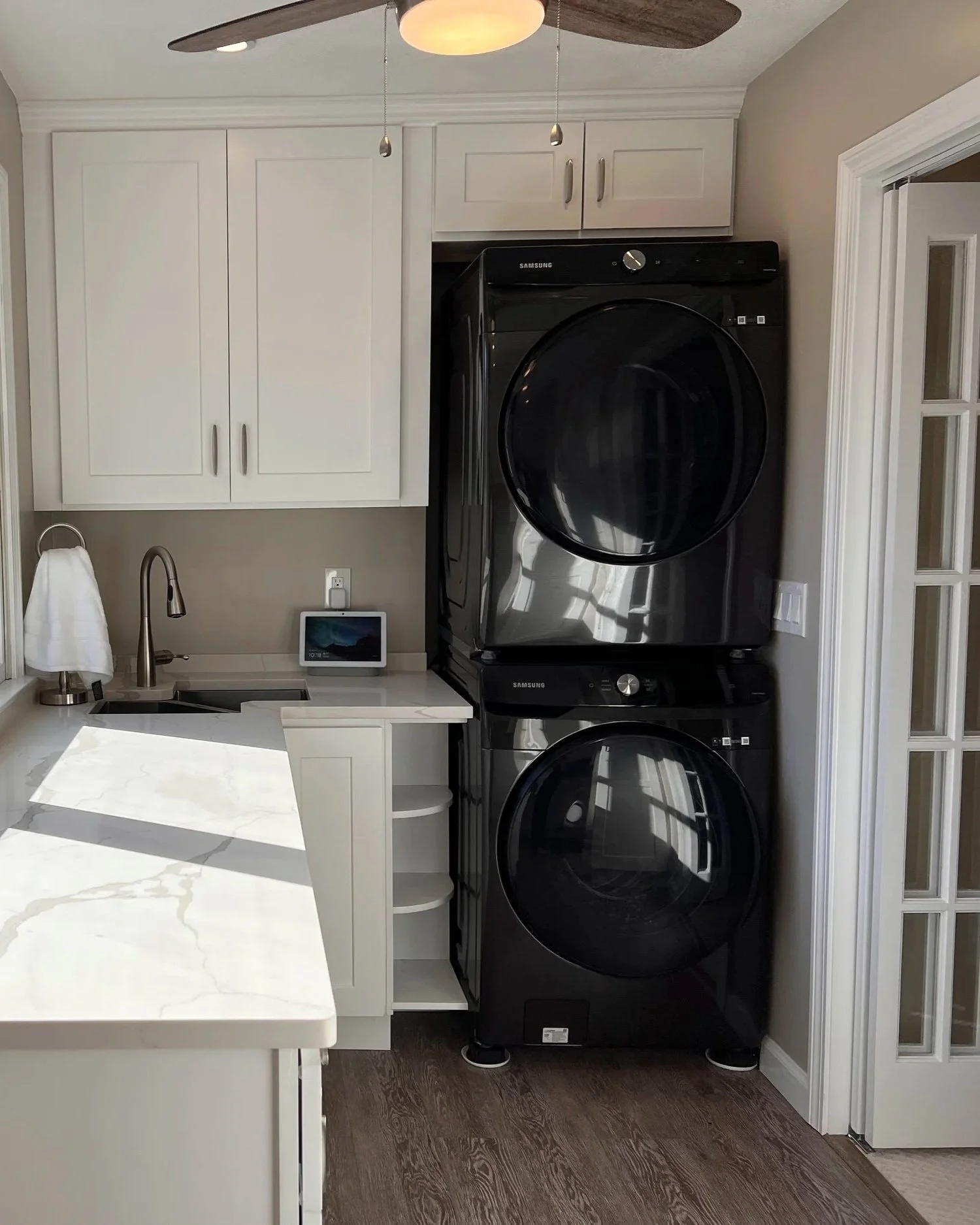 Stacked black Samsung washer and dryer in a laundry room with white cabinets, a small sink, a soap dispenser, and a smart home device on the countertop.