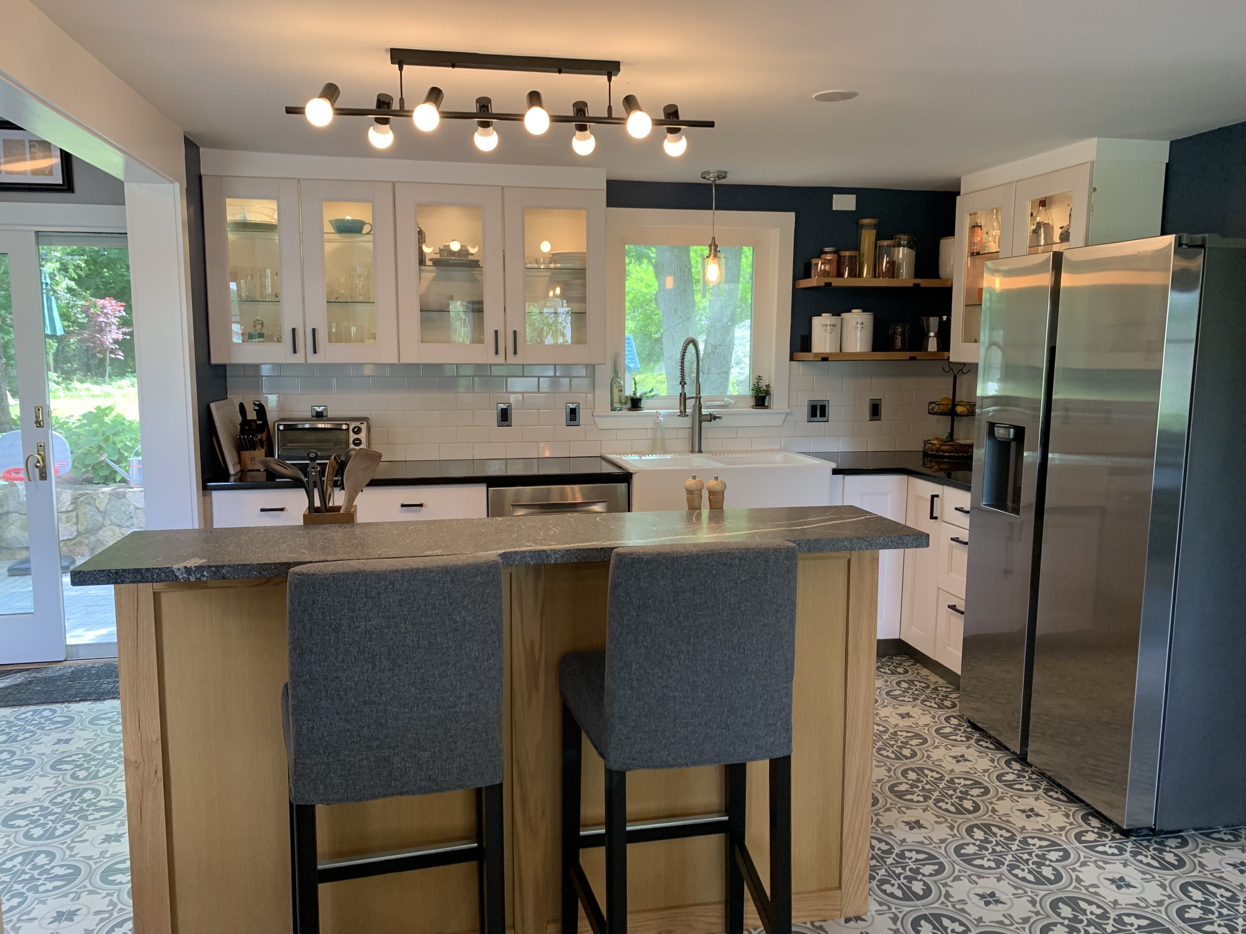 Modern kitchen with white cabinets, dark countertops, stainless steel appliances, patterned tile flooring, and a kitchen island with two dark gray chairs. There are hanging light fixtures, open shelves with jars, and a window showing greenery outside