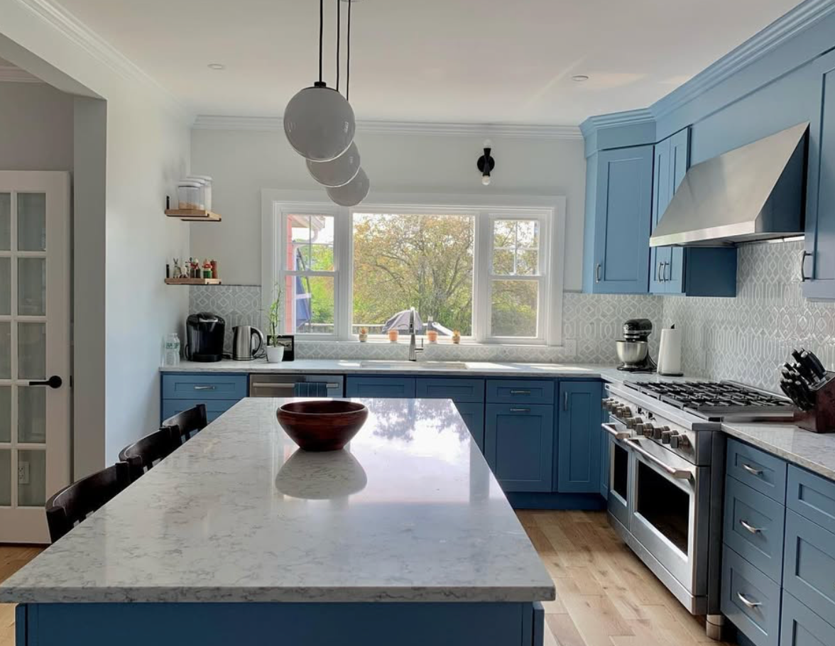 Modern kitchen with blue cabinets, stainless steel appliances, white marble countertop island, and a window overlooking trees.