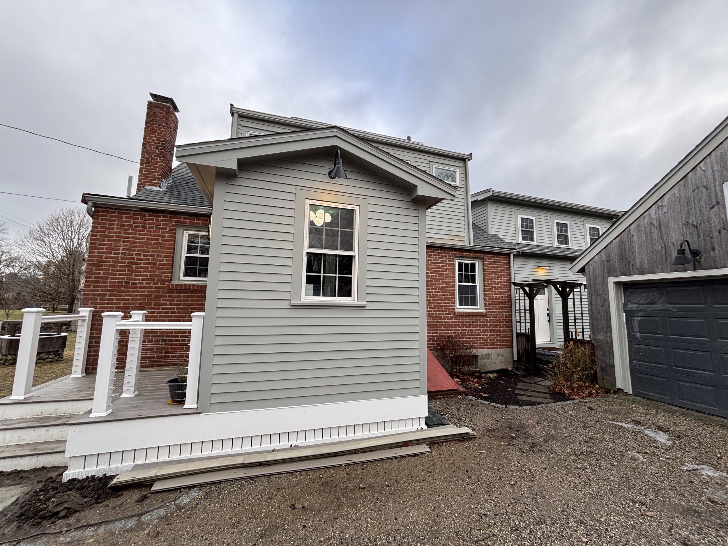 Exterior view of a house with gray siding and red brick walls, a small wooden porch with white railing, and a gravel driveway, under cloudy sky.