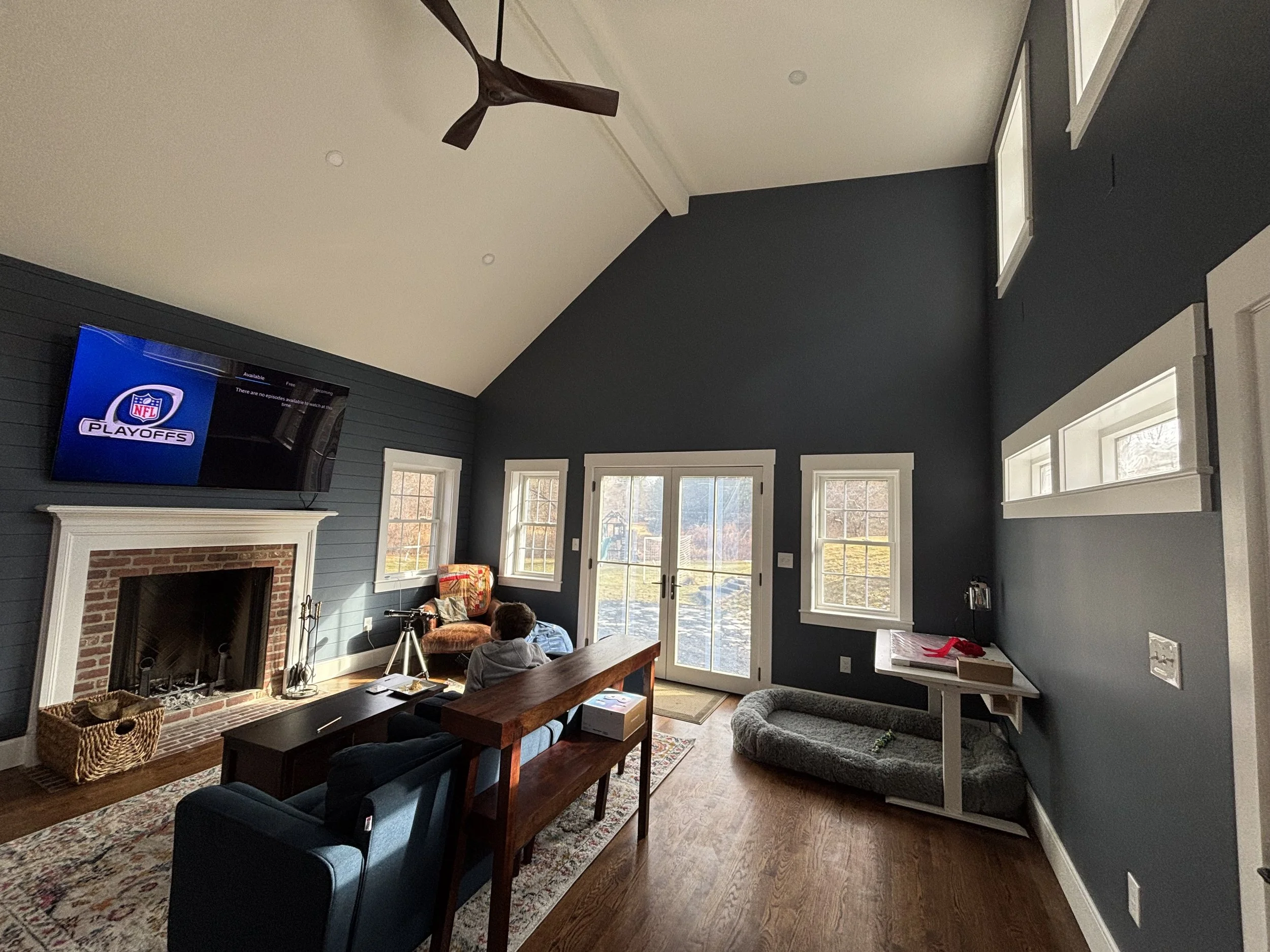 Living room with blue accent wall, fireplace, wall-mounted TV with NFL playoffs on screen, wooden coffee table, blue armchair, small child seated on sofa, large windows and glass doors letting in natural light, pet bed under windows, ceiling fan, har