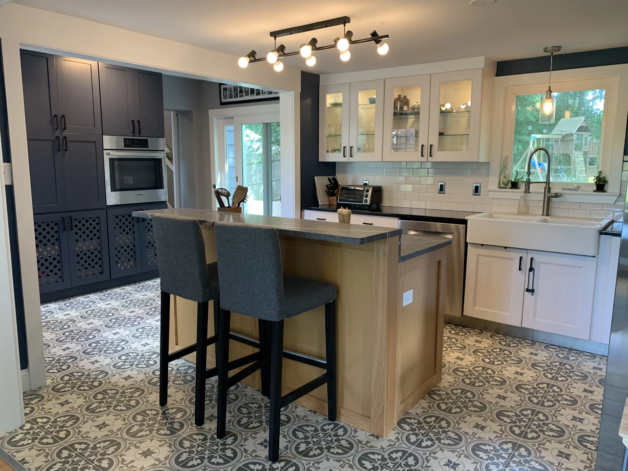 Modern kitchen with patterned tile floor, white and navy cabinetry, a central island with bar stools, white subway tile backsplash, stainless steel appliances, and a large window above the sink overlooking a backyard with playset.