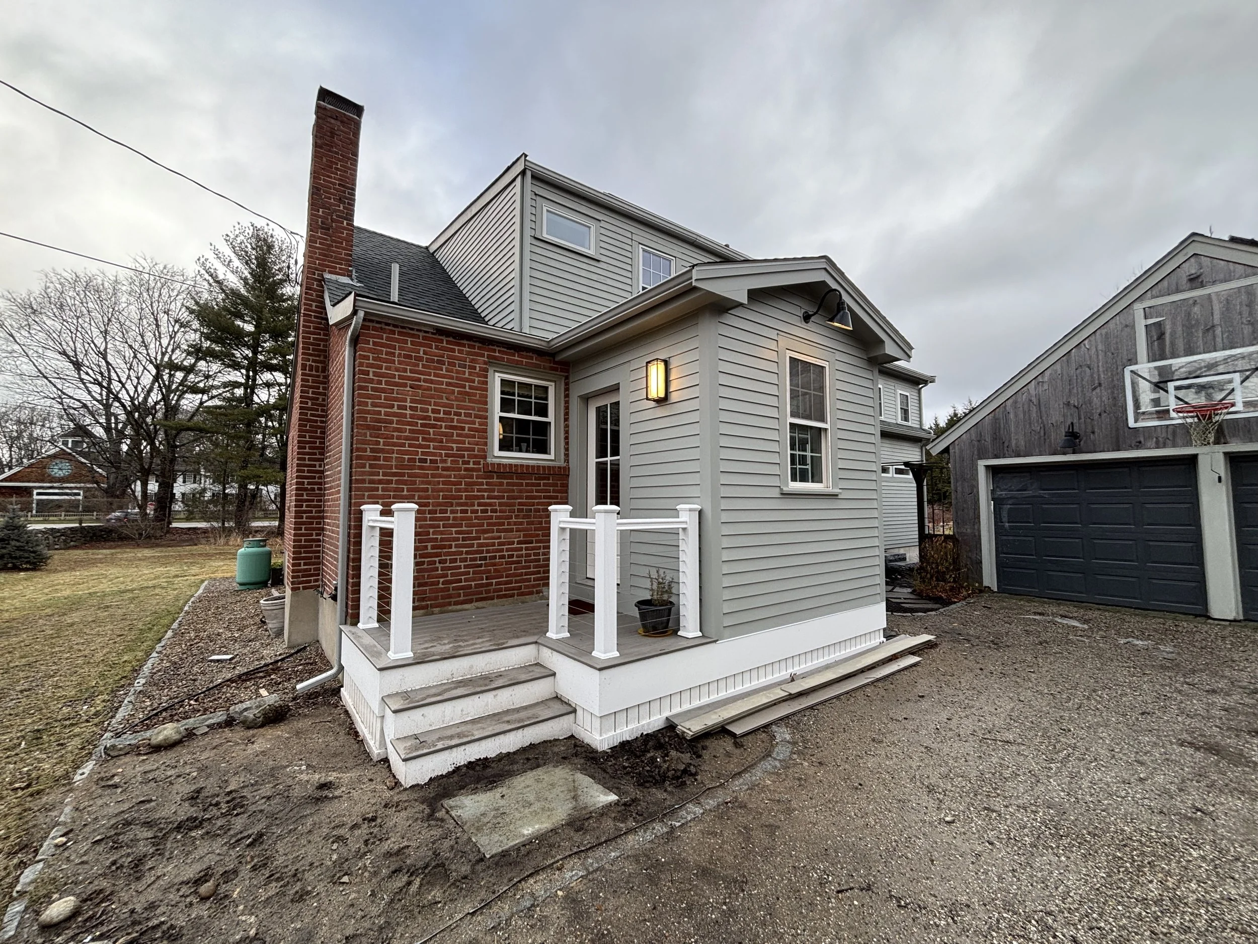 Backyard of a house with a small wooden porch, gray siding, a black outdoor light, and white railing, next to a brick wall with a chimney. A gravel driveway is in front, with a neighboring barn-like structure that has a basketball hoop above the gara