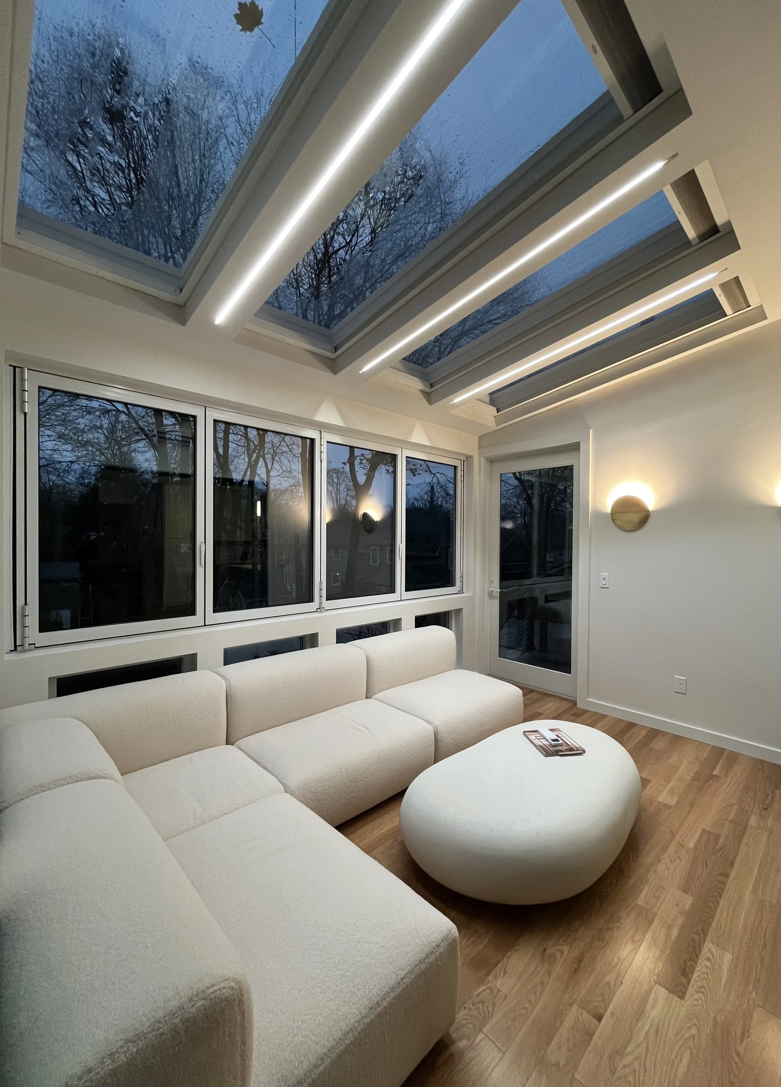 Modern living room with large skylights, white sectional sofa, round white ottoman, wood flooring, and wall-mounted lights.