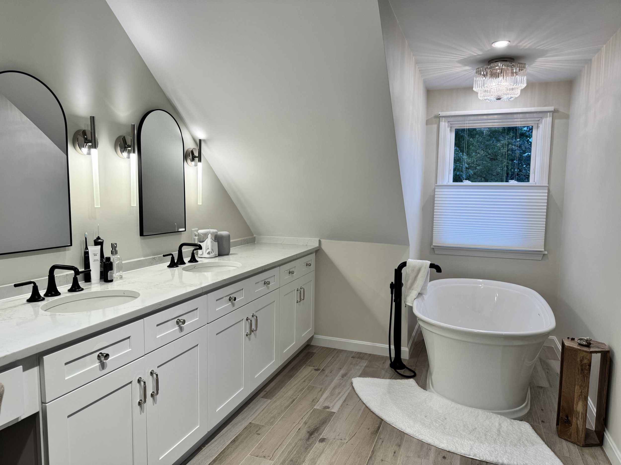 A modern bathroom with a white double vanity, black faucets, three mirrors, and wall-mounted lights. There is a freestanding tub with a white towel on a black towel rack, a window with a view of trees, and a small wooden table next to the tub.