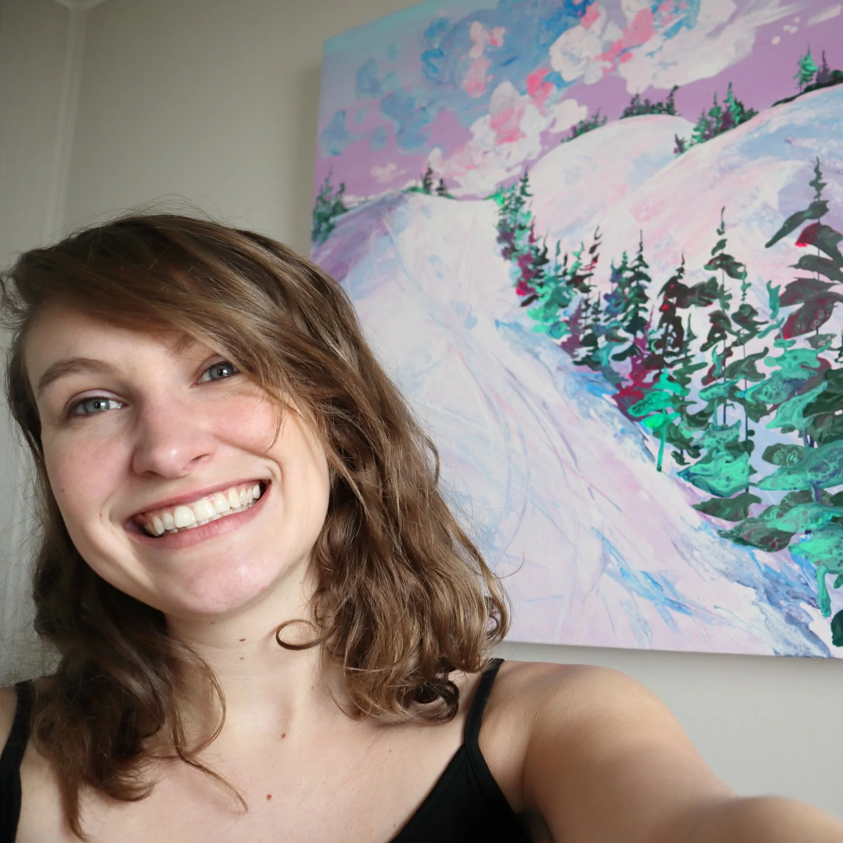 A woman with wavy brown hair smiling takes a selfie in front of an abstract painting of a snow-covered landscape with trees.