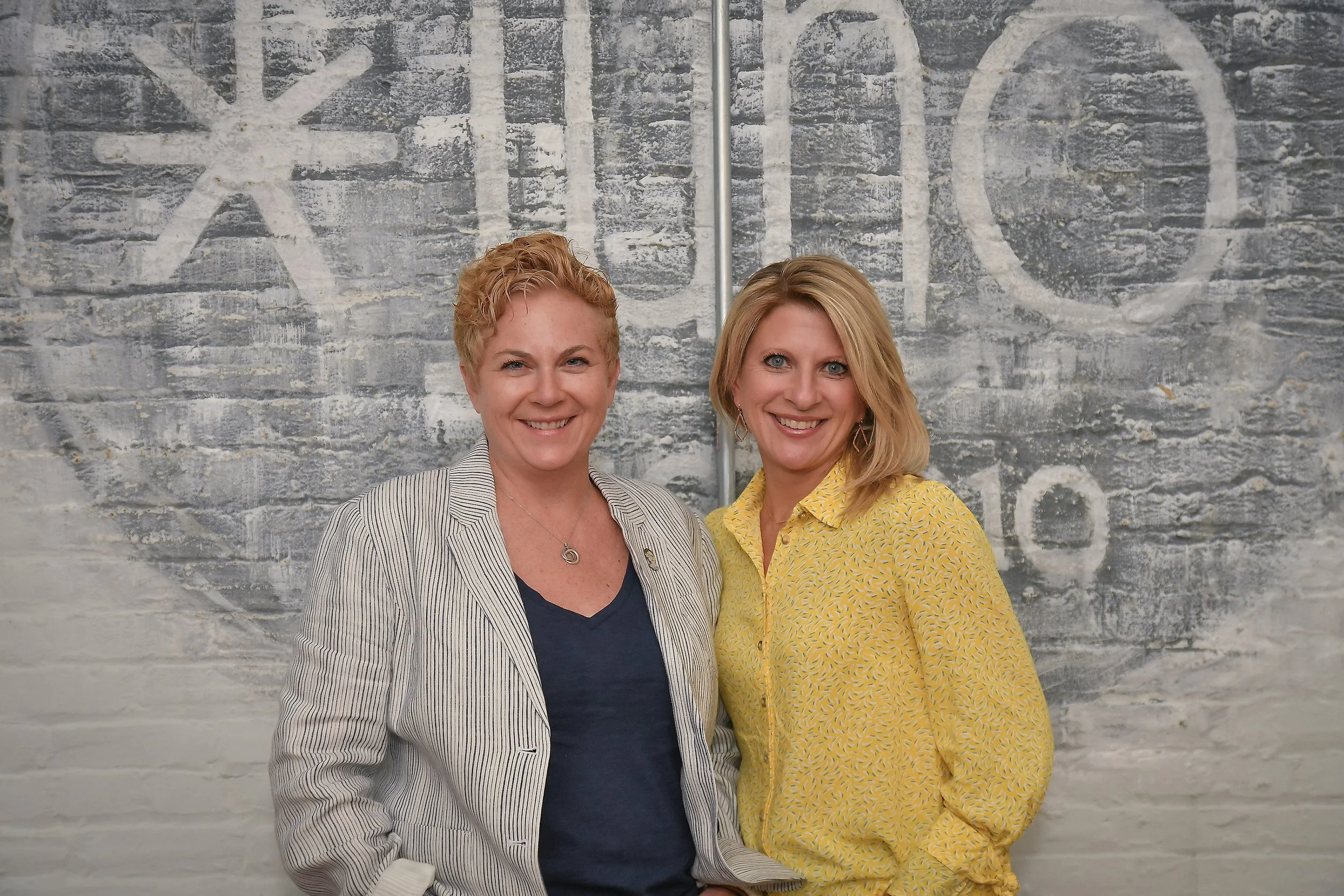 Photo of Mikal Harden and Vicki Sack in business casual clothes standing in front of a white brick wall with a gray Juno logo painted on it