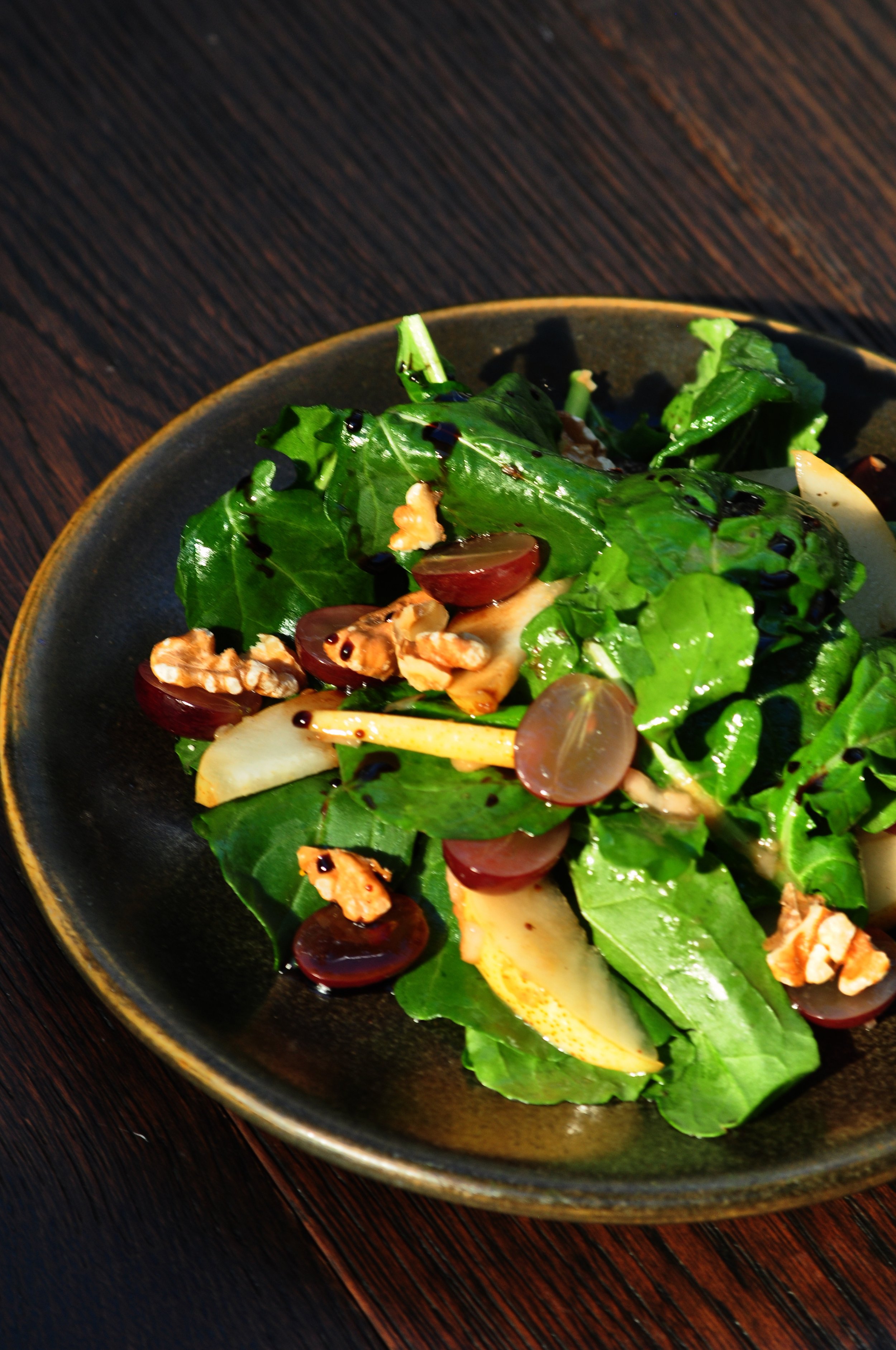 Fresh mixed green salad with grapes, walnuts, and dressing in a black bowl on a wooden table.